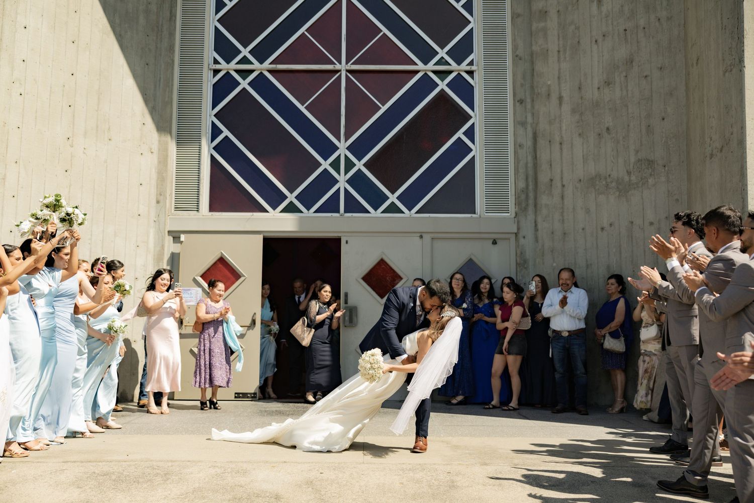 Bride and groom share romantic dip kiss outside church, surrounded by cheering bridesmaids and wedding guests.