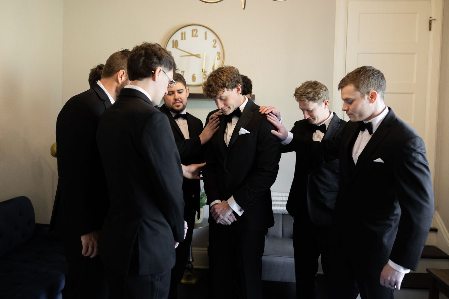 Groomsmen in black tuxedos gather together before a wedding ceremony, sharing a moment near a vintage wall clock.