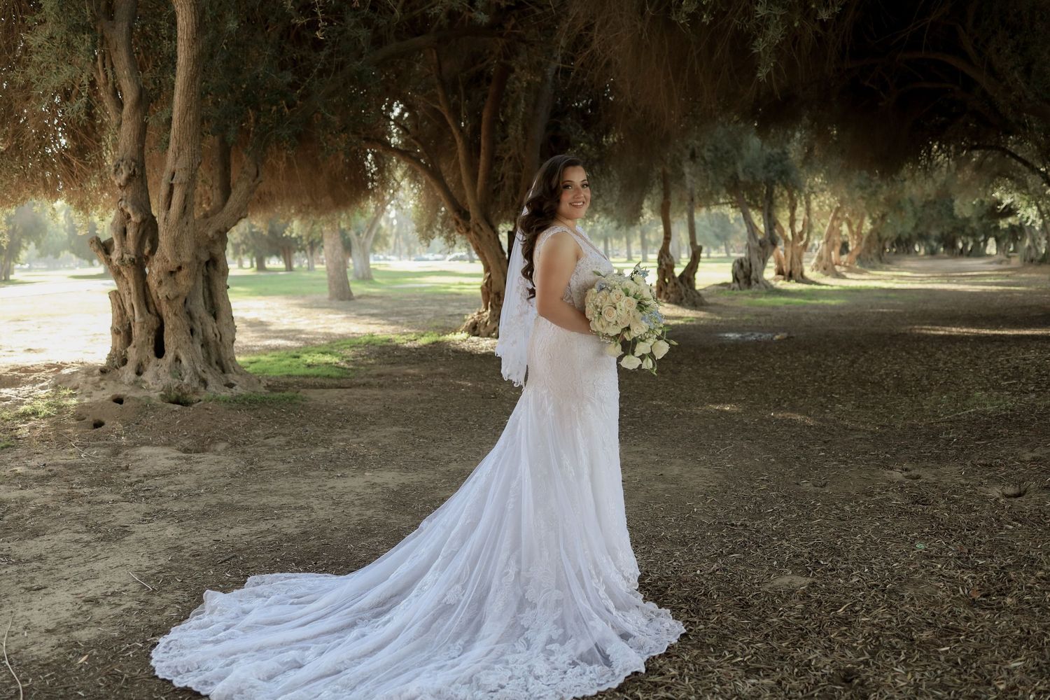 Bride in flowing white lace gown poses among majestic trees in outdoor setting.