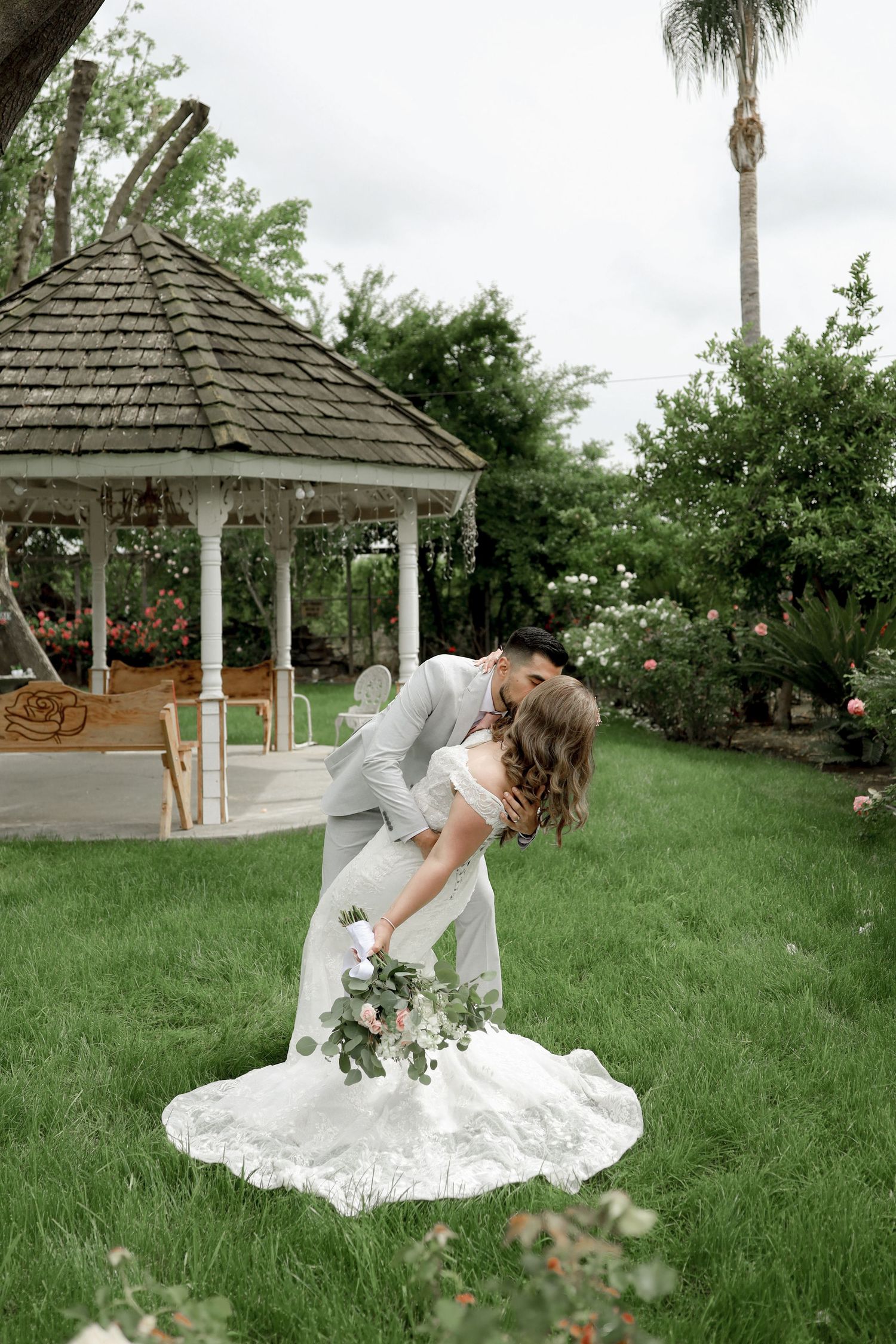 White lace wedding dress spread out on green grass near a garden gazebo.