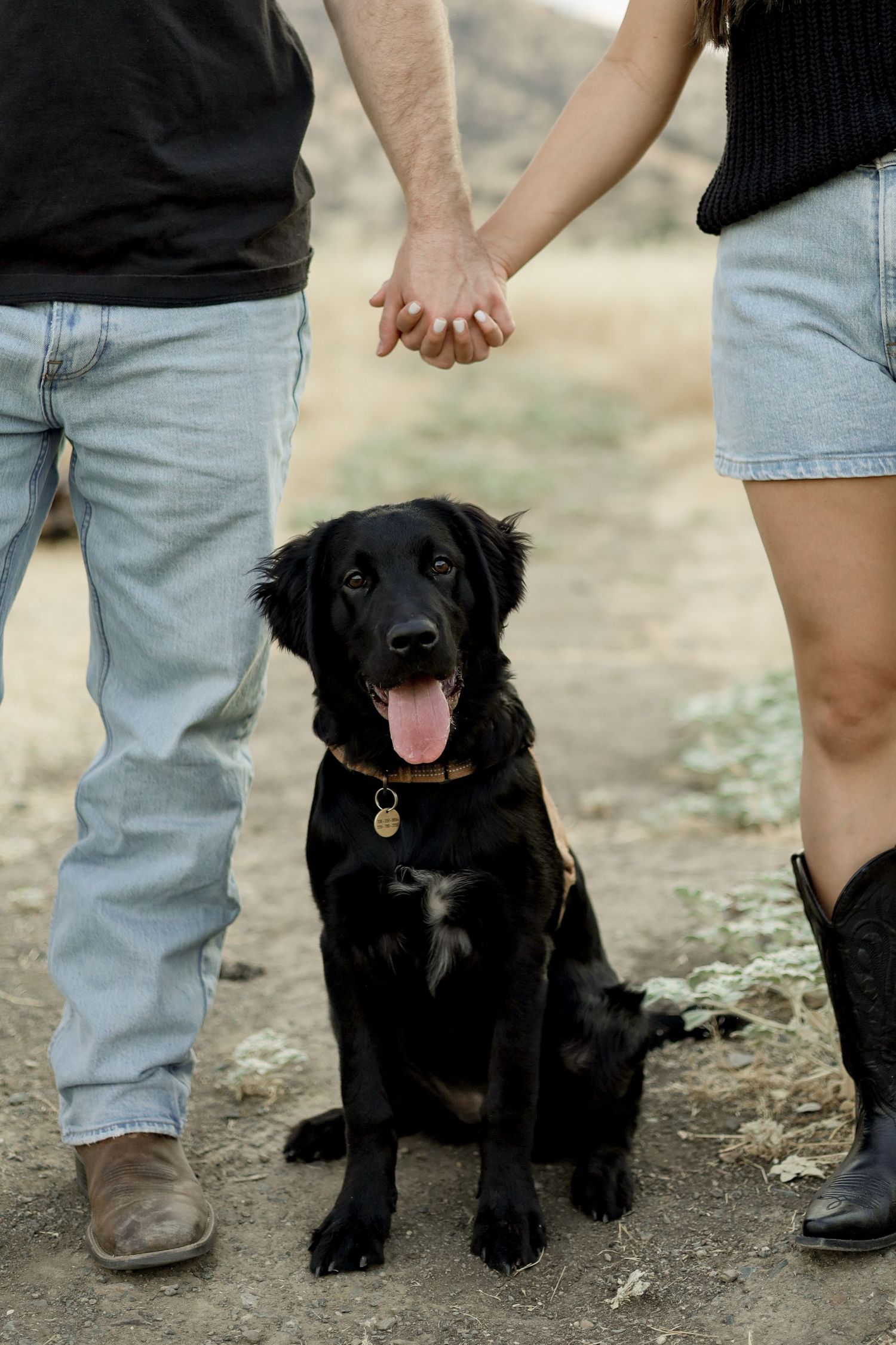 Happy black dog sitting between two people wearing casual attire on a dirt path.