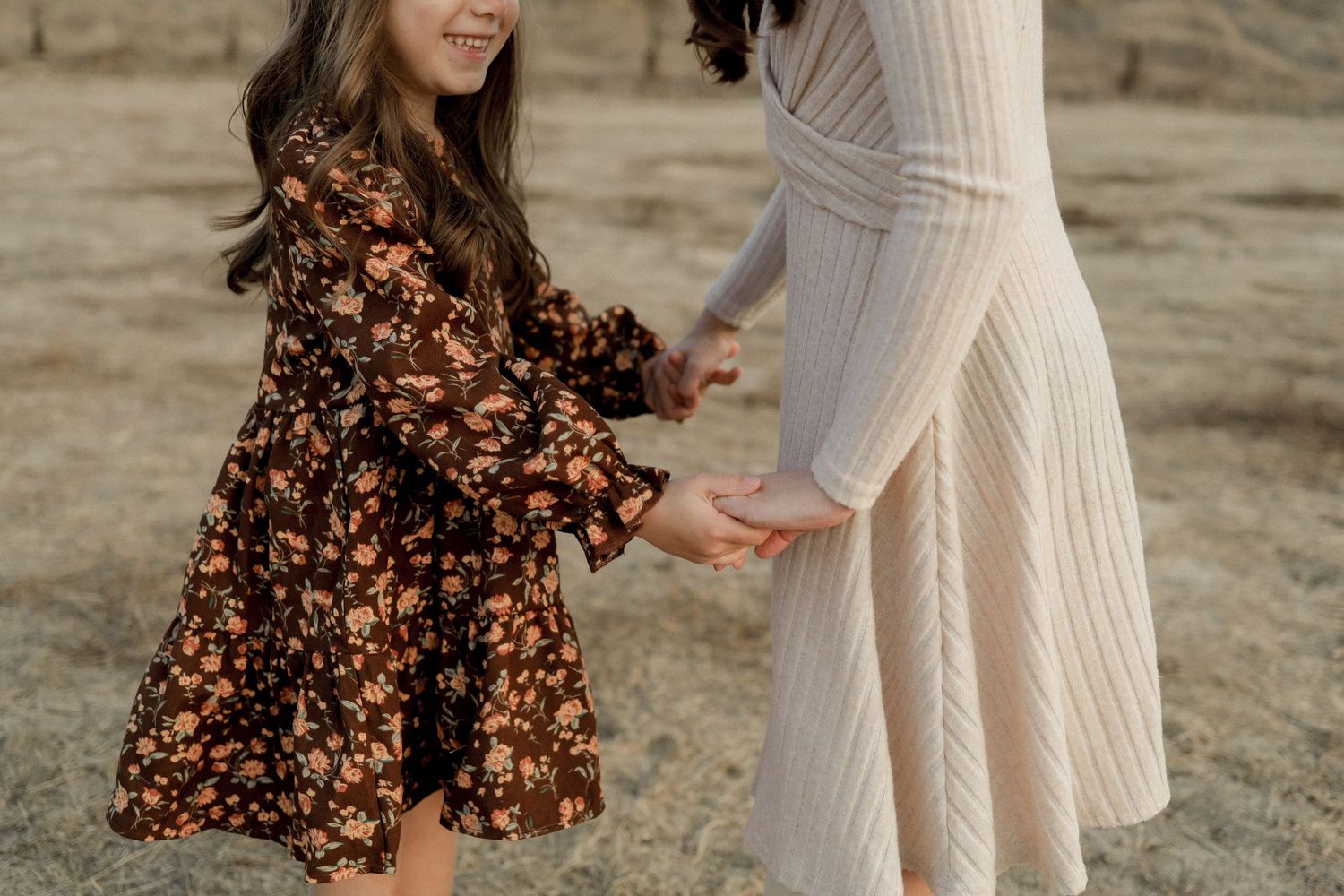 A woman in a white dress and brown floral dress twirl and dance together outdoors.