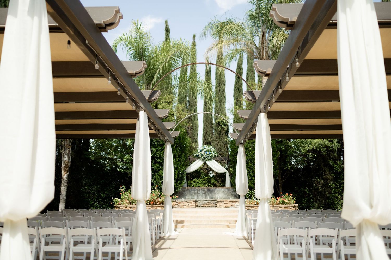 Outdoor wedding ceremony setup with white drapes and chairs lined up under palm trees.