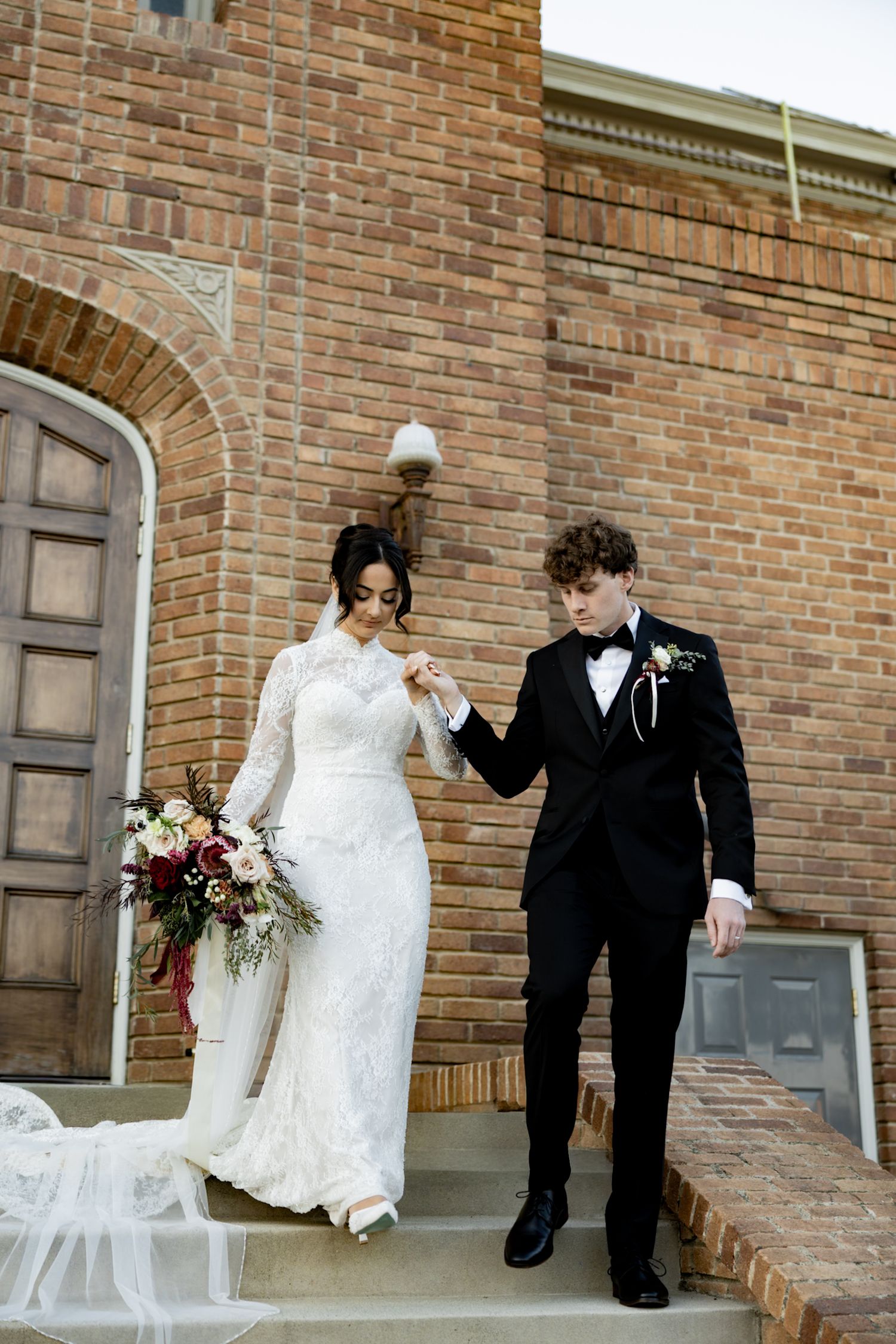 Bride in lace gown holds bouquet as groom in tuxedo holds her hand on church steps.