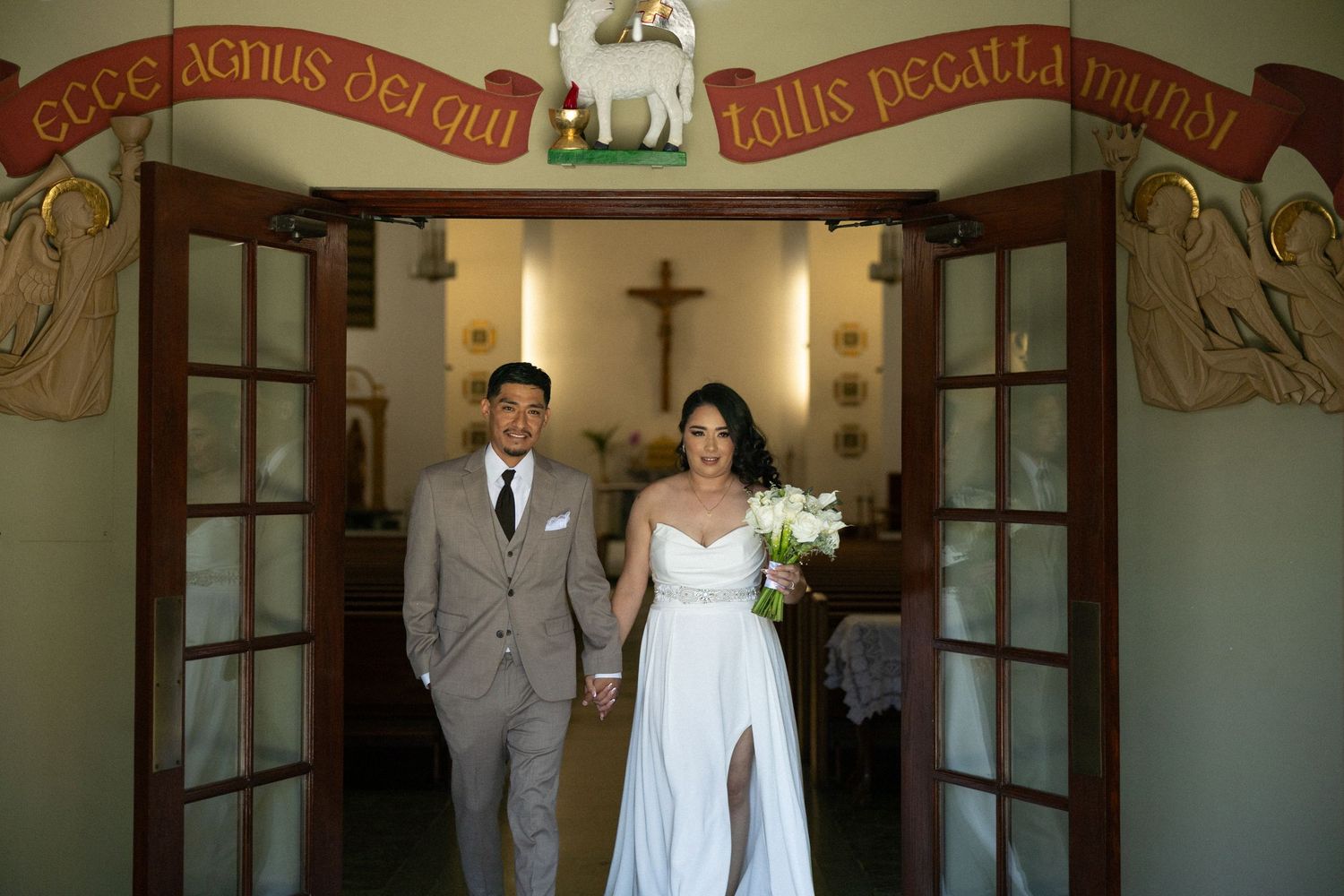 Bride and groom exit a chapel doorway holding hands, with Latin inscription banner above the French doors.