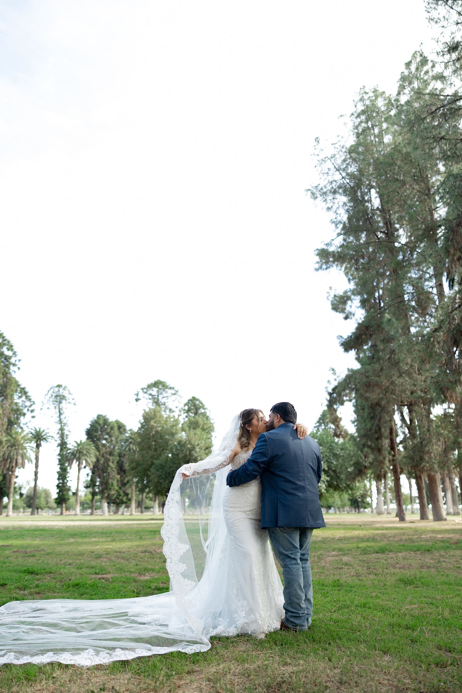 Bride and groom share a romantic kiss outdoors, her long white veil flowing across green grass surrounded by tall trees.