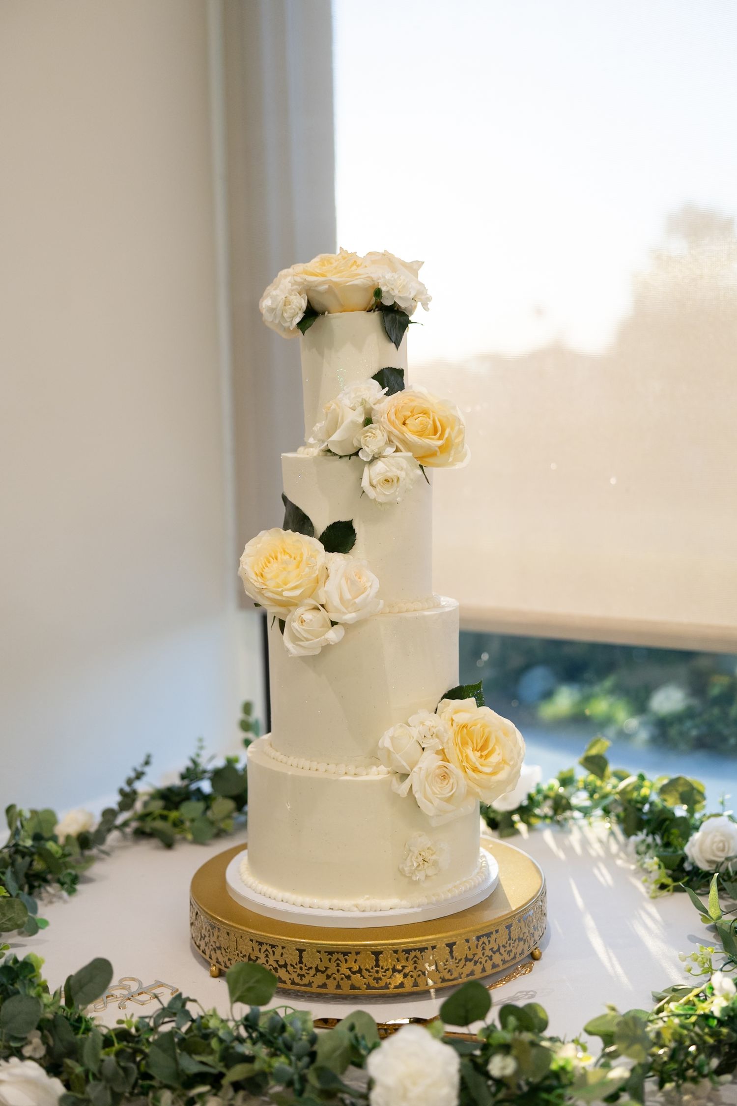 Four-tier white wedding cake adorned with cream roses and greenery displayed on gold cake stand by window.