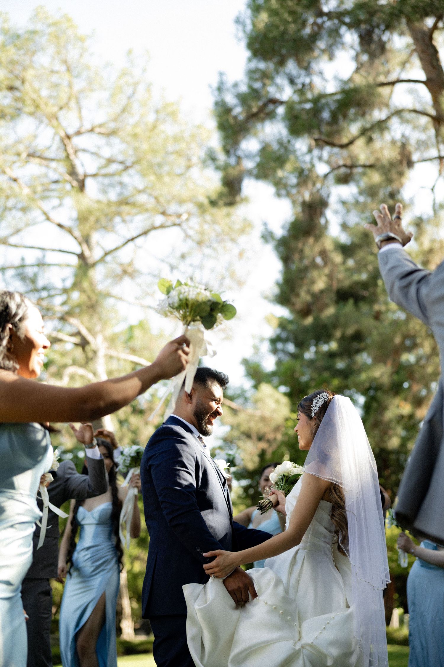 Bride and groom walk through cheering guests outdoors at a sunny garden wedding ceremony.