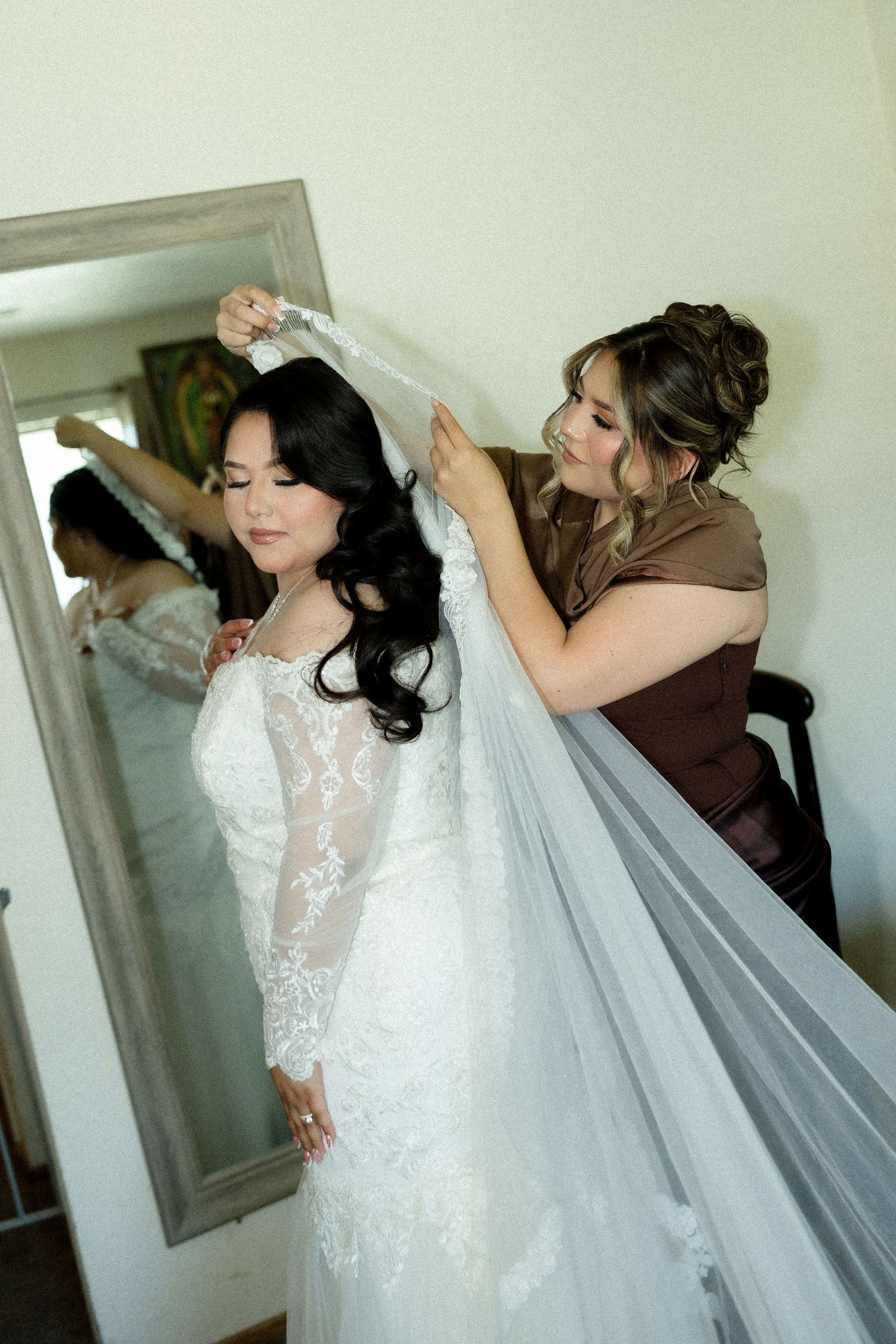 A bridesmaid in brown helps a bride in lace wedding gown put on her veil in front of a mirror.