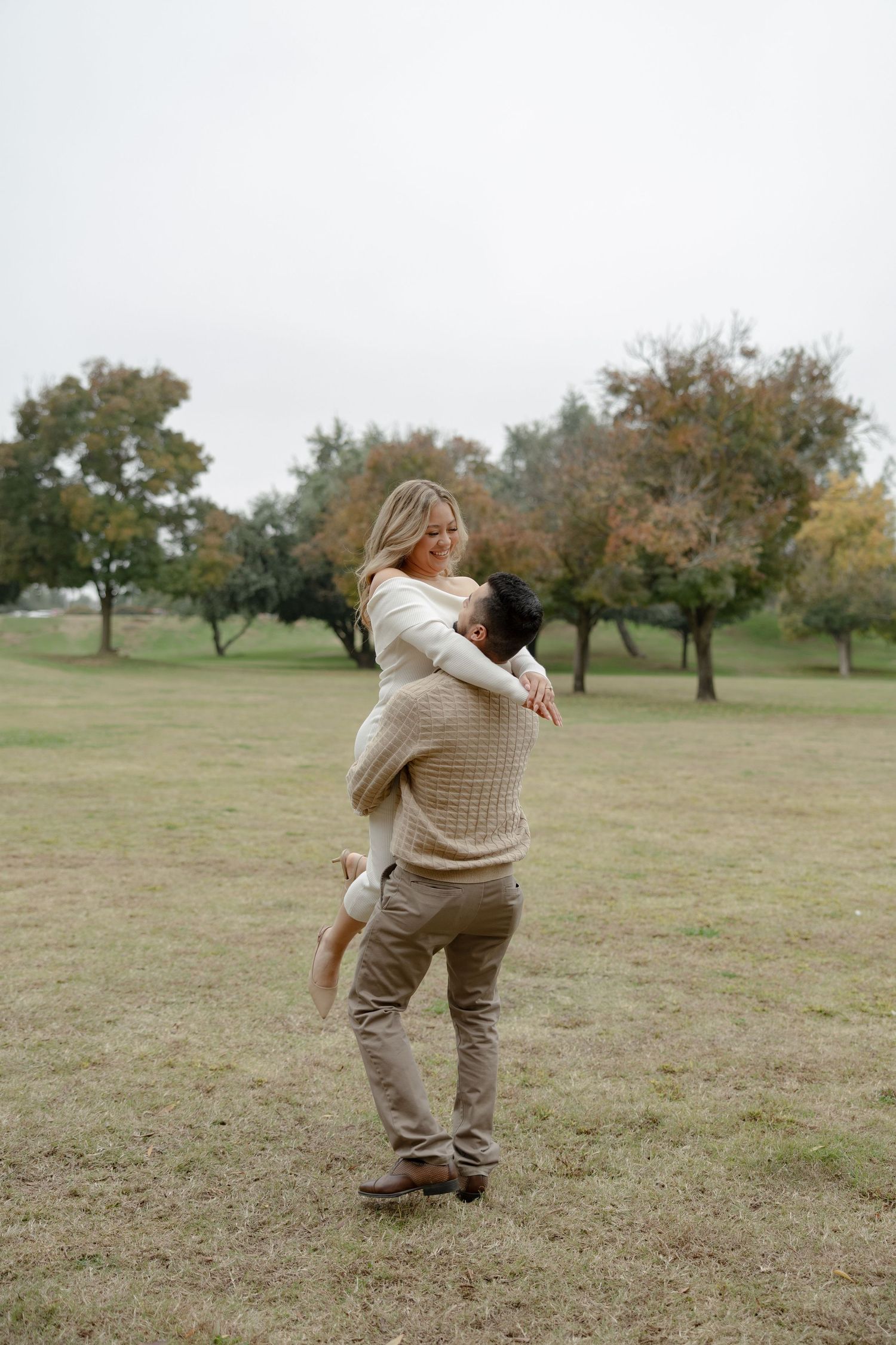 Two people in beige outfits dancing and embracing playfully in a grassy field with autumn trees.