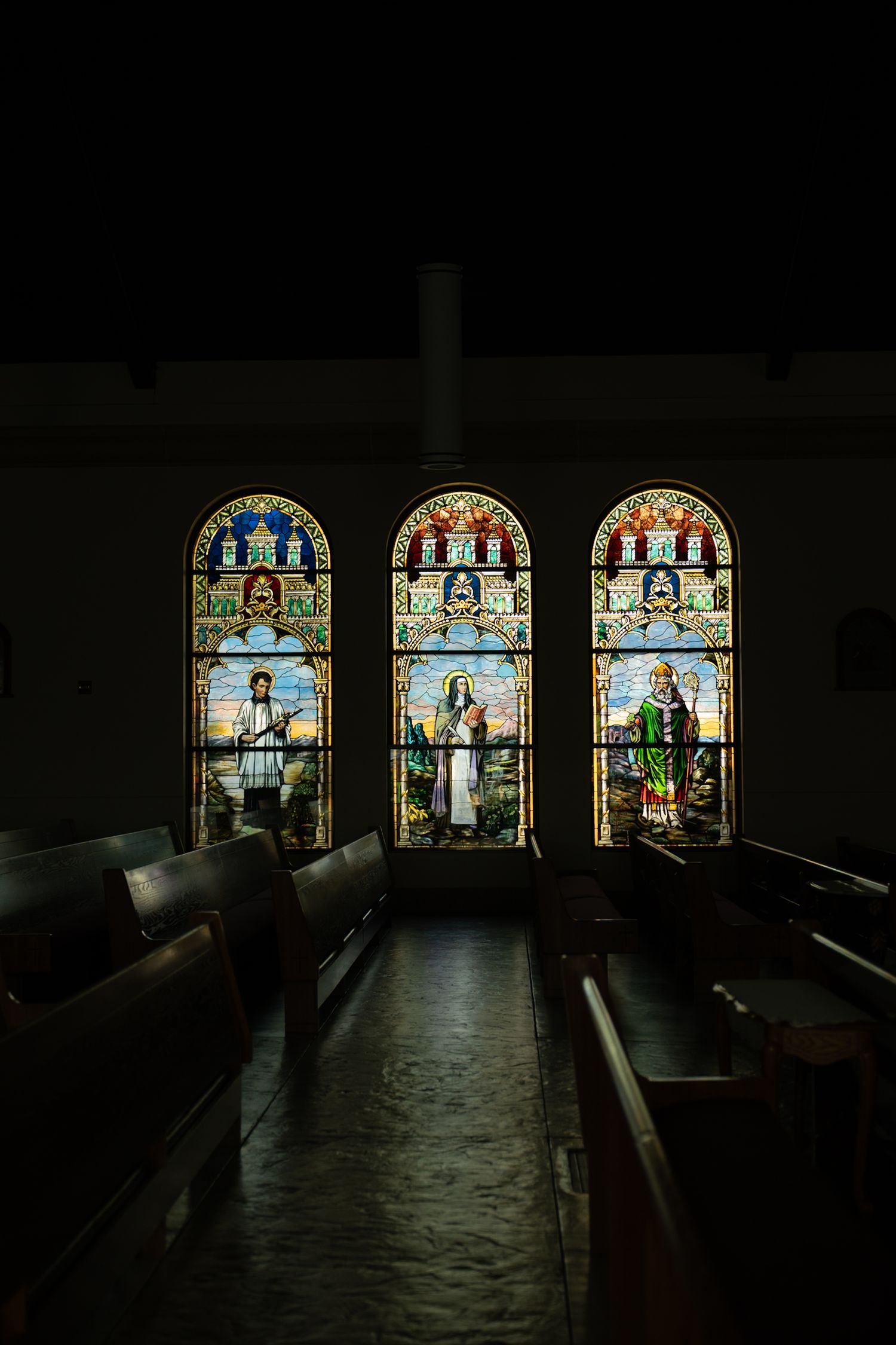 Dark church interior with illuminated stained glass windows casting colorful light on wooden pews.