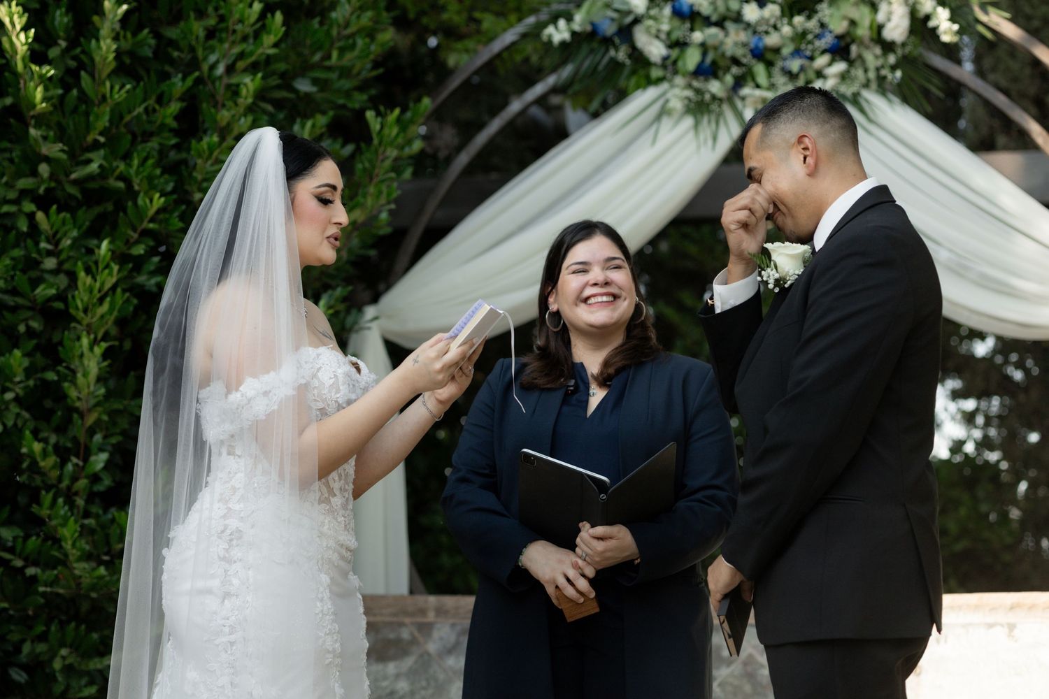 A couple exchanges rings during an outdoor wedding ceremony under a white canopy surrounded by greenery.
