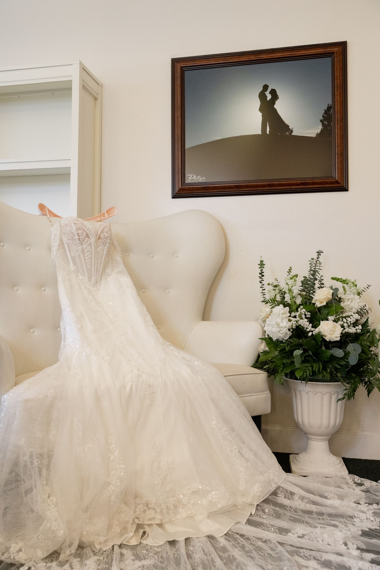Elegant lace wedding dress draped over white tufted chair with bridal bouquet and romantic silhouette artwork above.