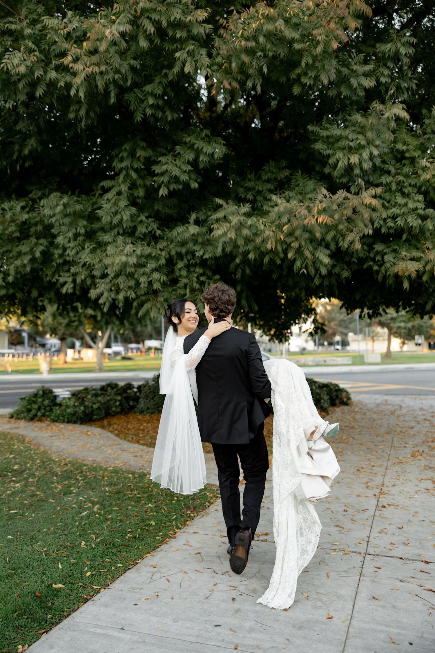 Bride and groom walking on sidewalk near large green trees, bride in white gown with train, groom in black suit.
