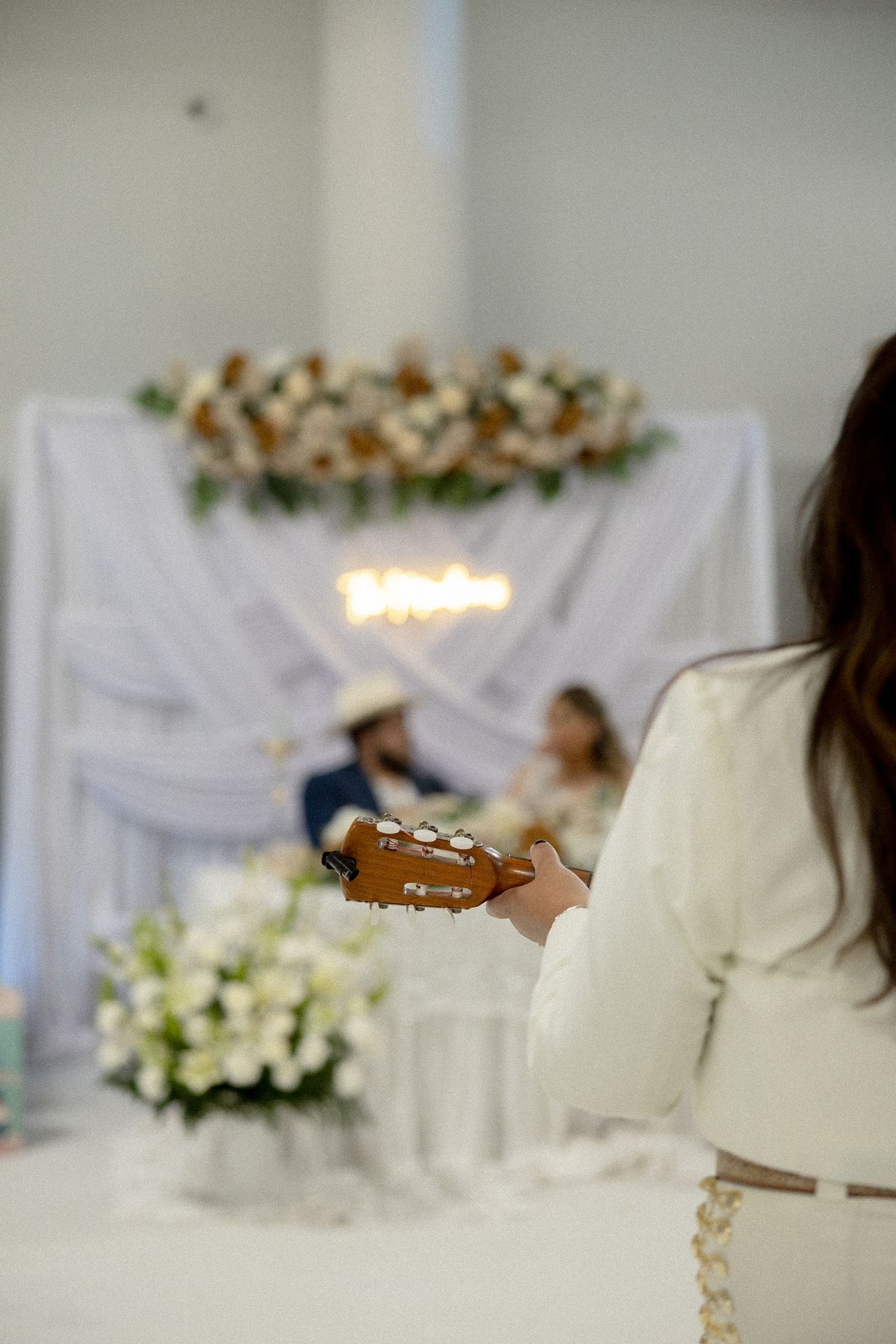 Musician playing guitar at elegant wedding ceremony with floral arch backdrop and couple seated in background.