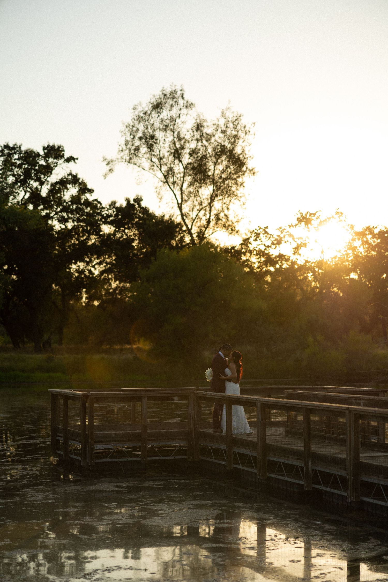 Couple sharing a romantic kiss on a wooden bridge at golden sunset, surrounded by lush trees reflected in calm water.