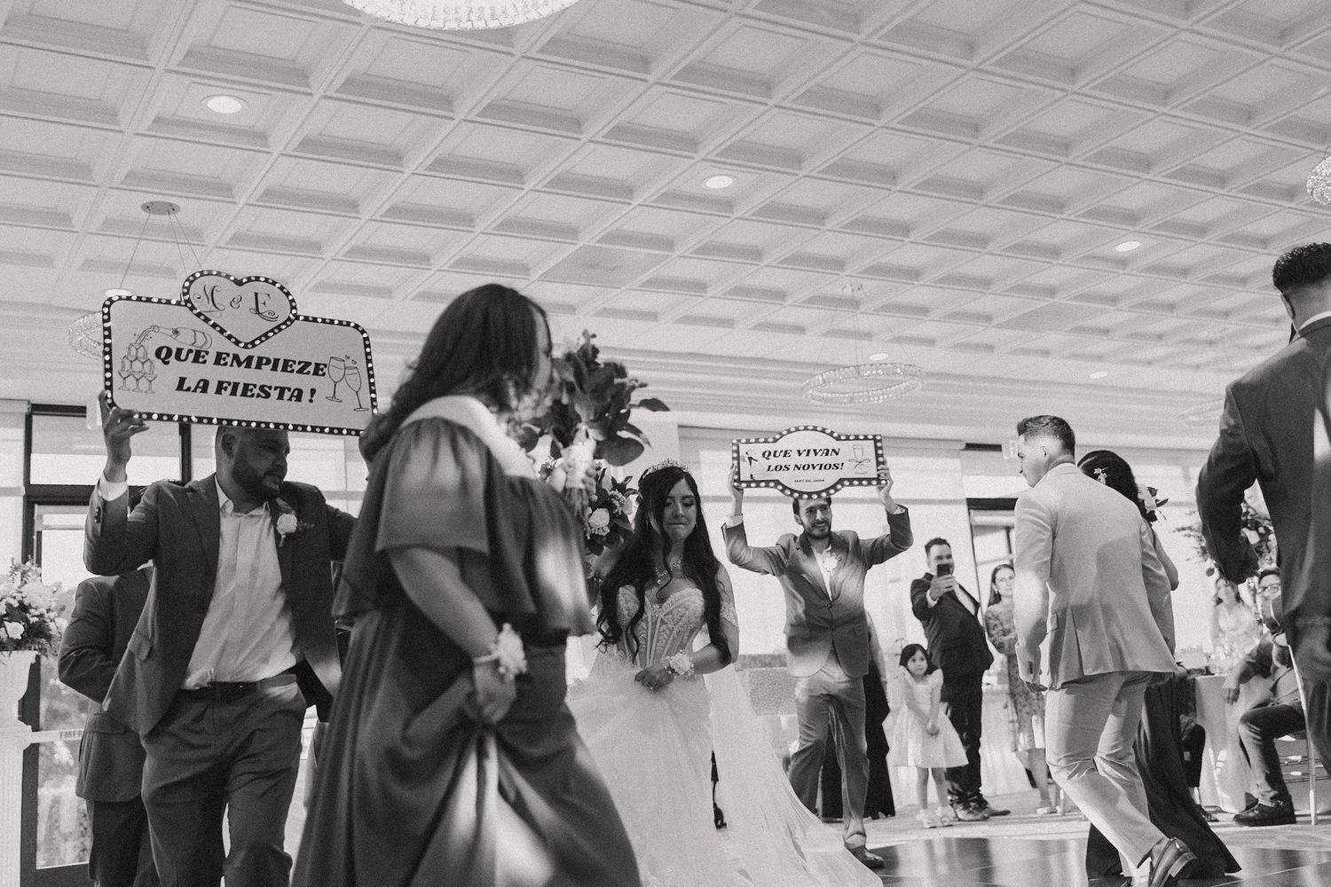 Black and white wedding photo with guests holding signs reading Que Empiece La Fiesta during a festive celebration.