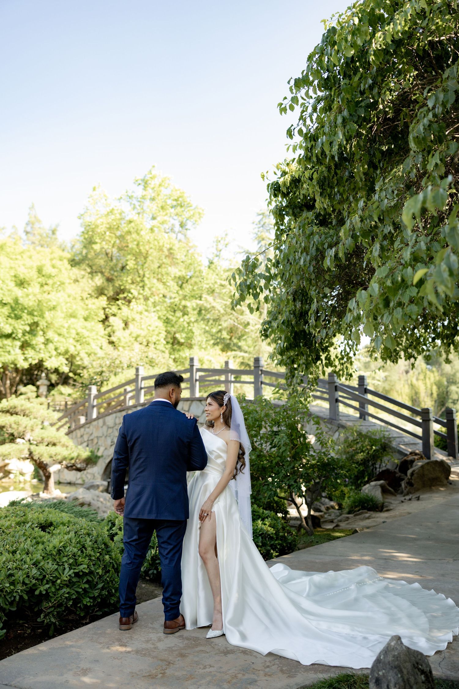 Bride in white gown and veil stands with groom in navy suit on garden path near wooden bridge.