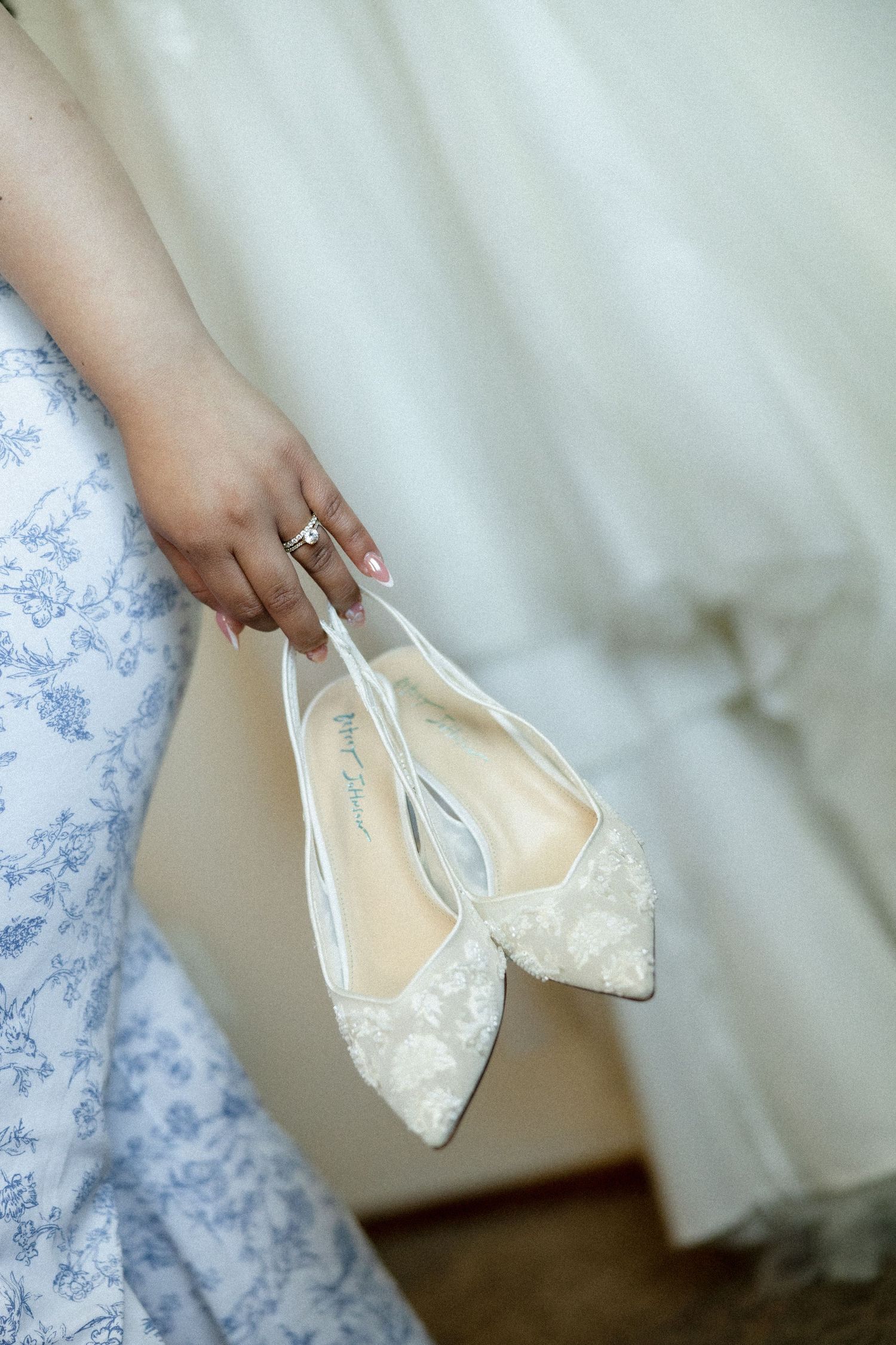 A bride holds elegant white lace pointed-toe flat wedding shoes with bow detail, wearing a floral blue dress and diamond ring.