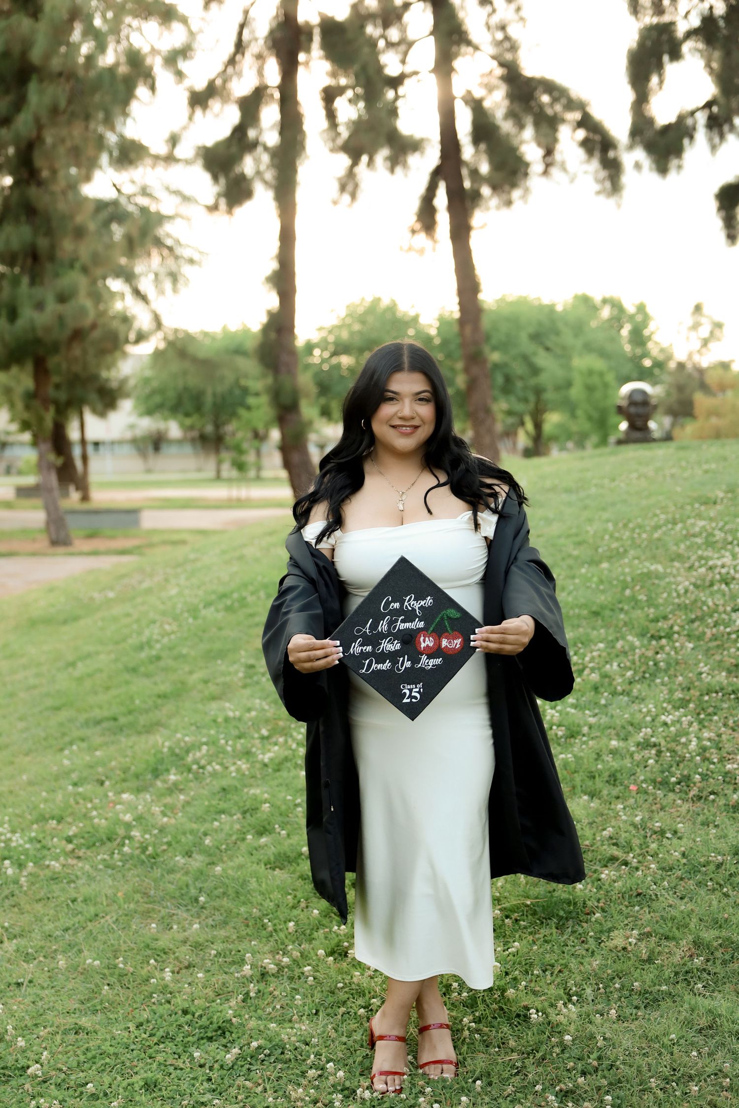 Graduate in white dress and black graduation gown holds decorated cap while standing on grass near trees at sunset.