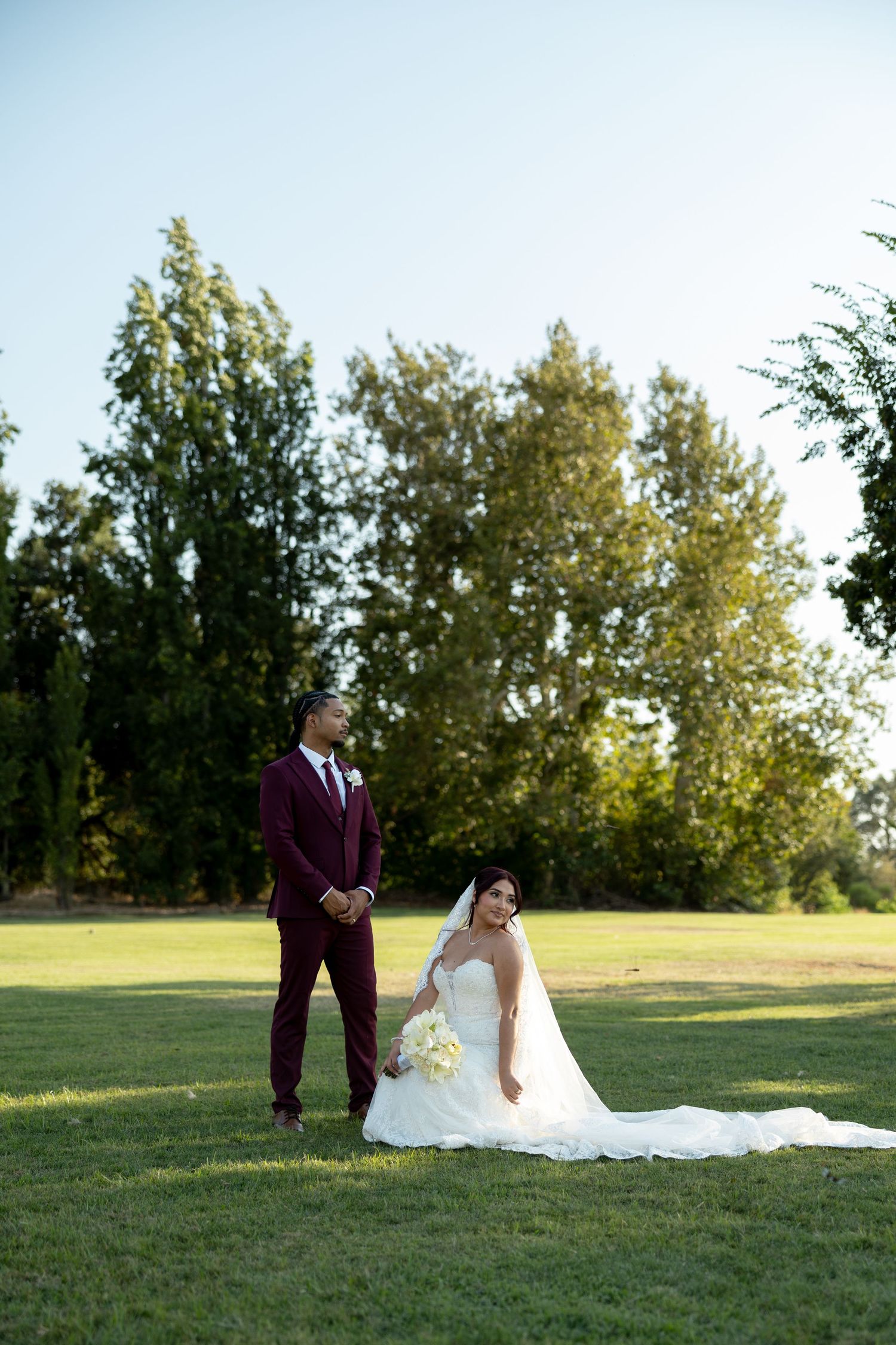 Groom in burgundy suit stands beside bride in white gown sitting gracefully on green lawn.