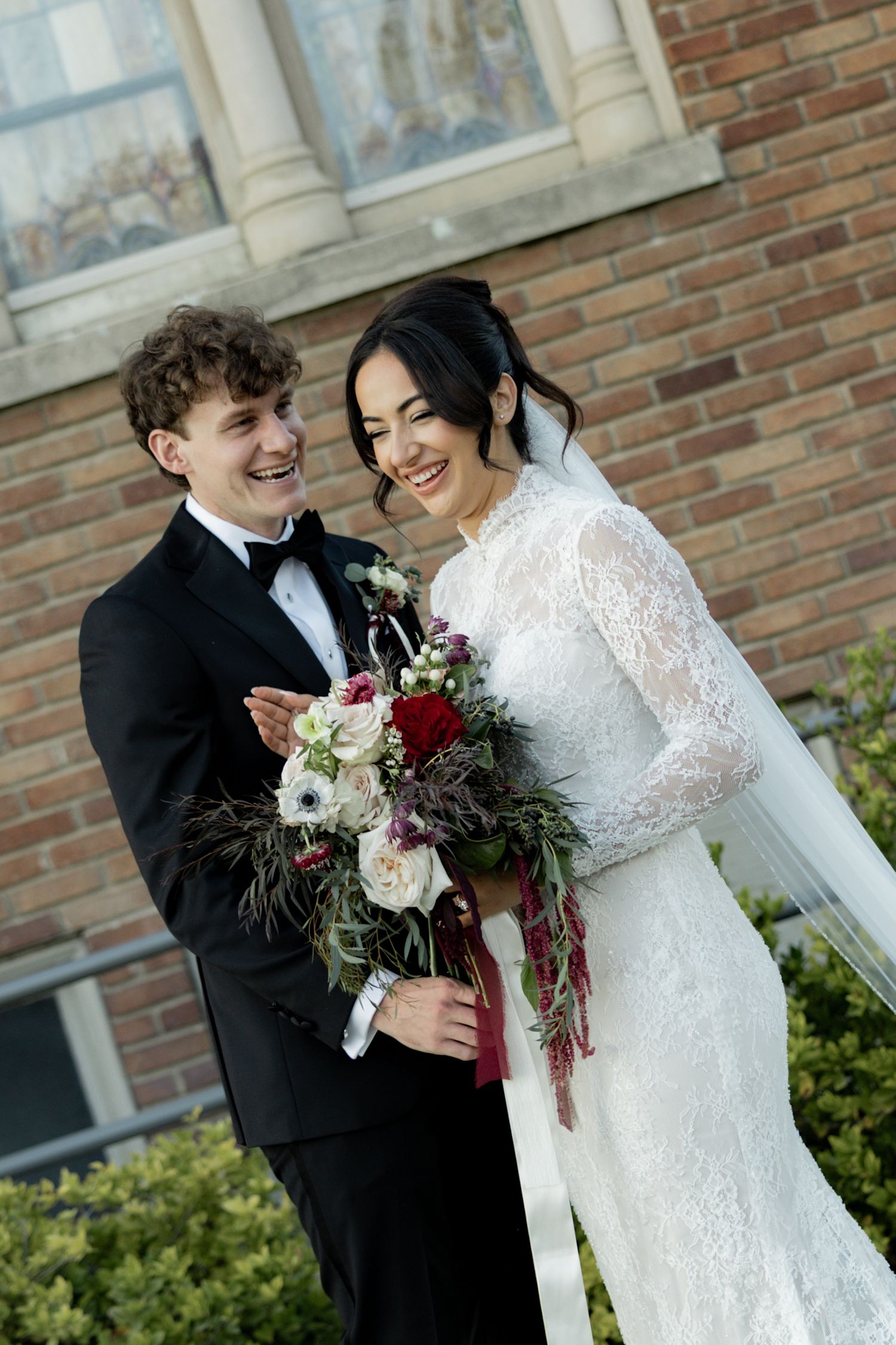 Smiling bride and groom pose outside brick church, bride holds burgundy and white floral bouquet.