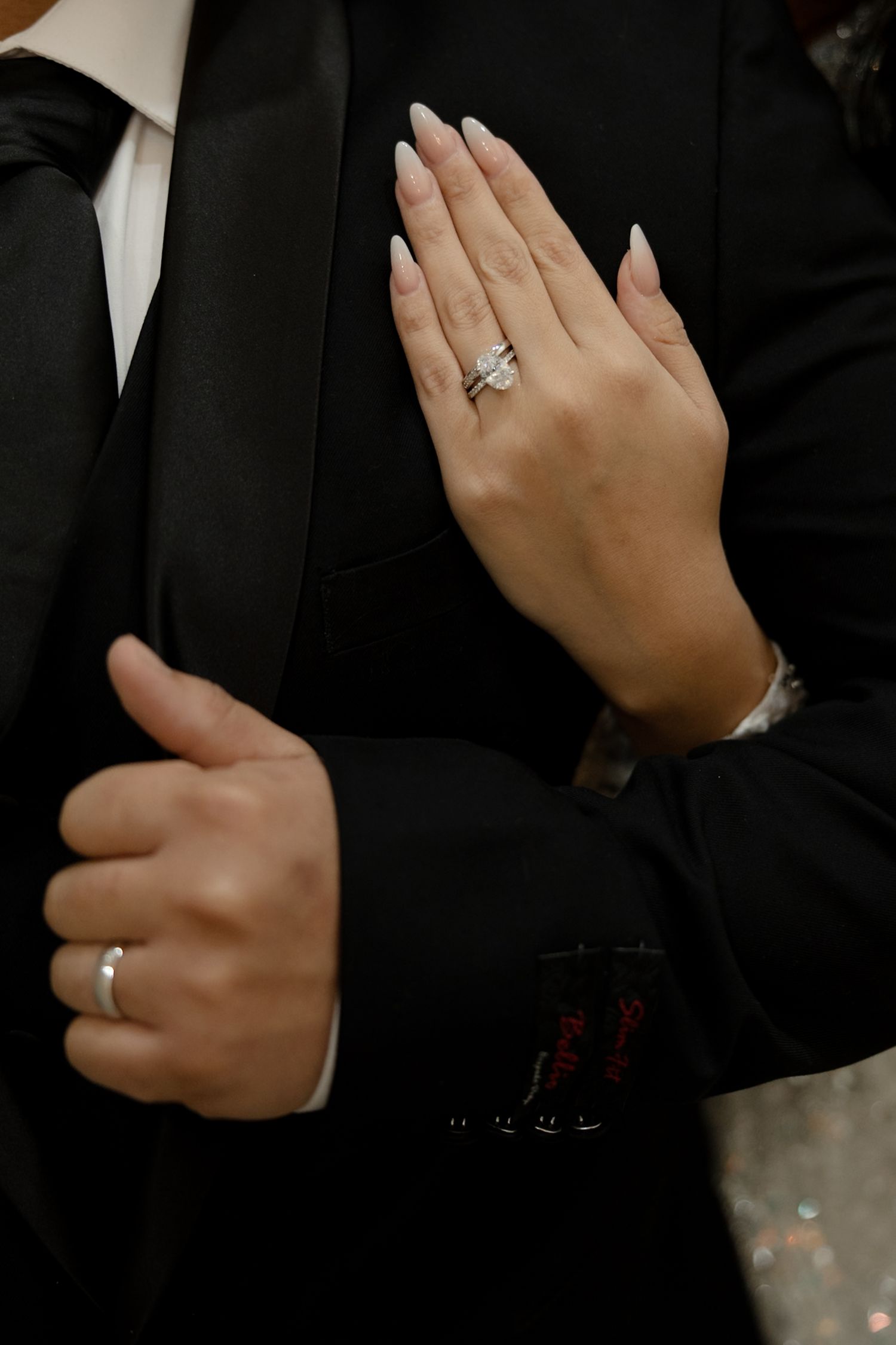 Close-up of hands showing diamond engagement ring against black formal attire.