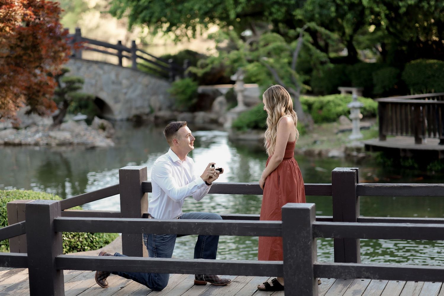 A couple shares a romantic proposal moment on a wooden bridge in a scenic garden setting.
