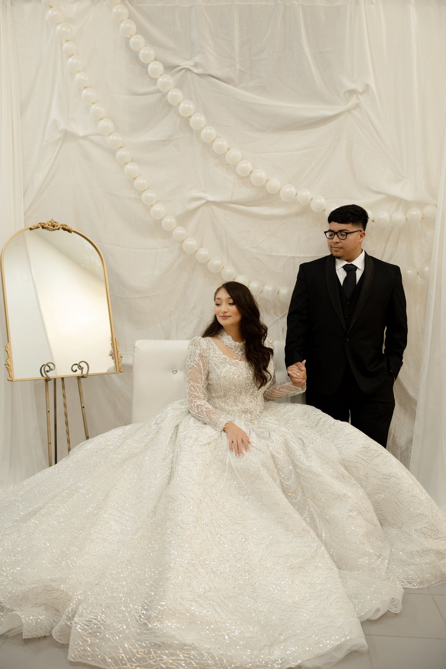 Elegant wedding photoshoot with couple in formal attire against white pearl-decorated backdrop.