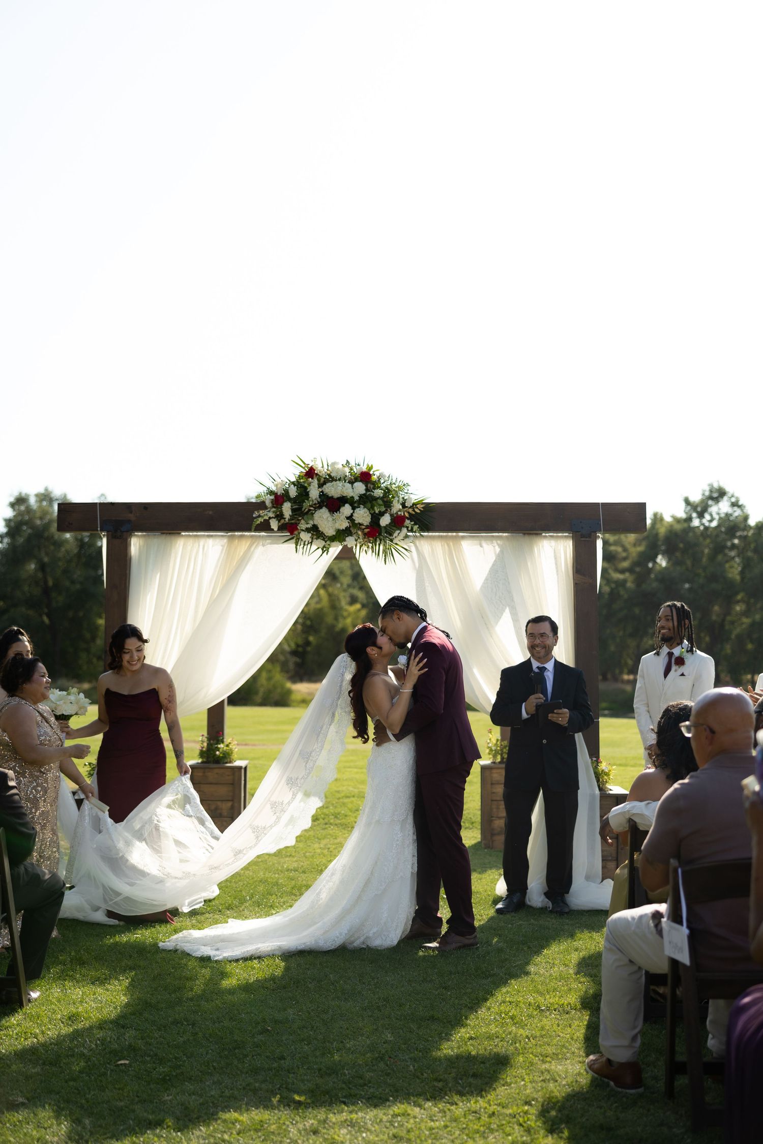 Bride and groom share first kiss at outdoor wedding ceremony under floral arch with white draping on green lawn.