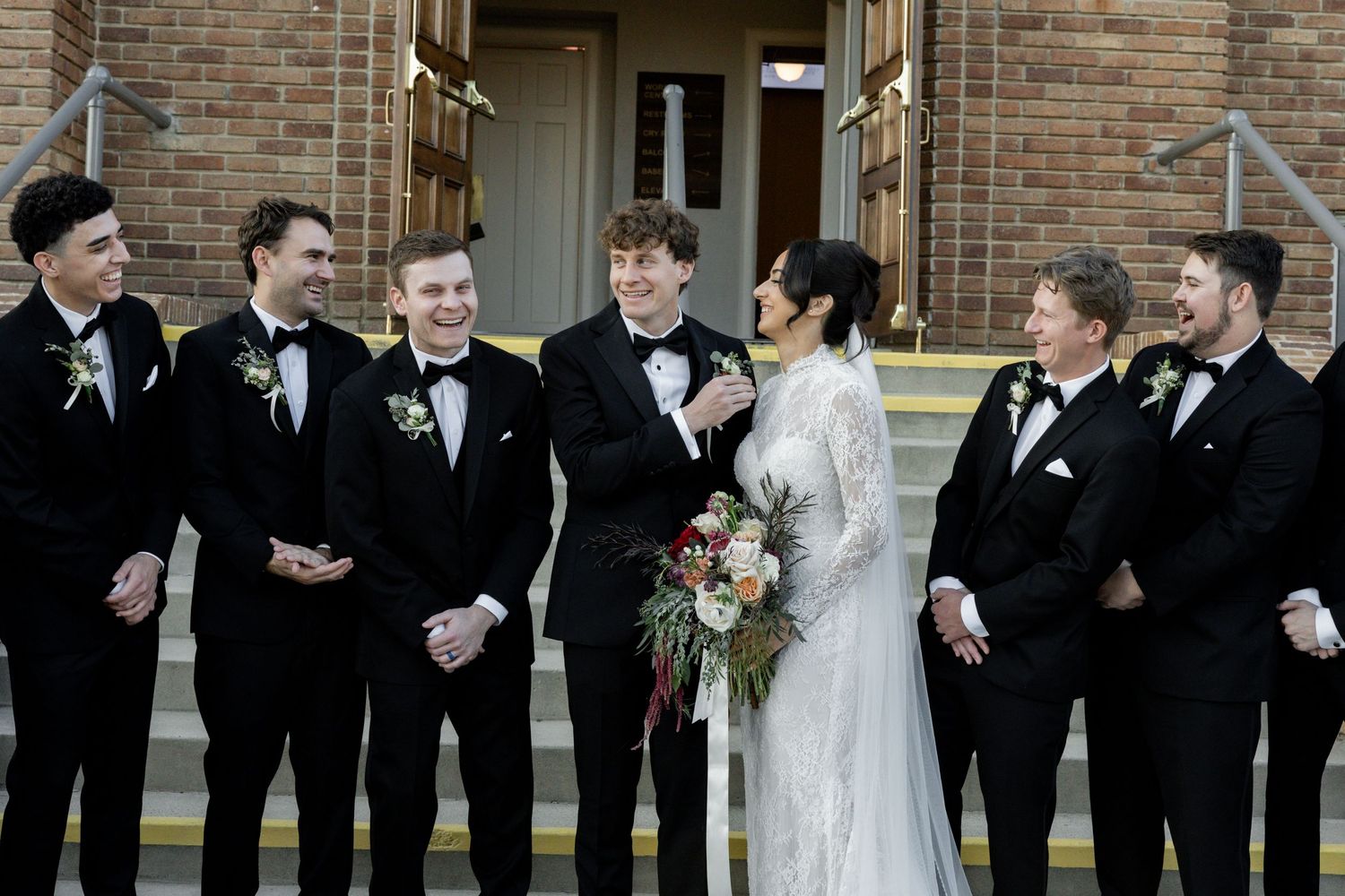 Bride in white lace gown holds bouquet surrounded by groomsmen in black tuxedos outside brick church.