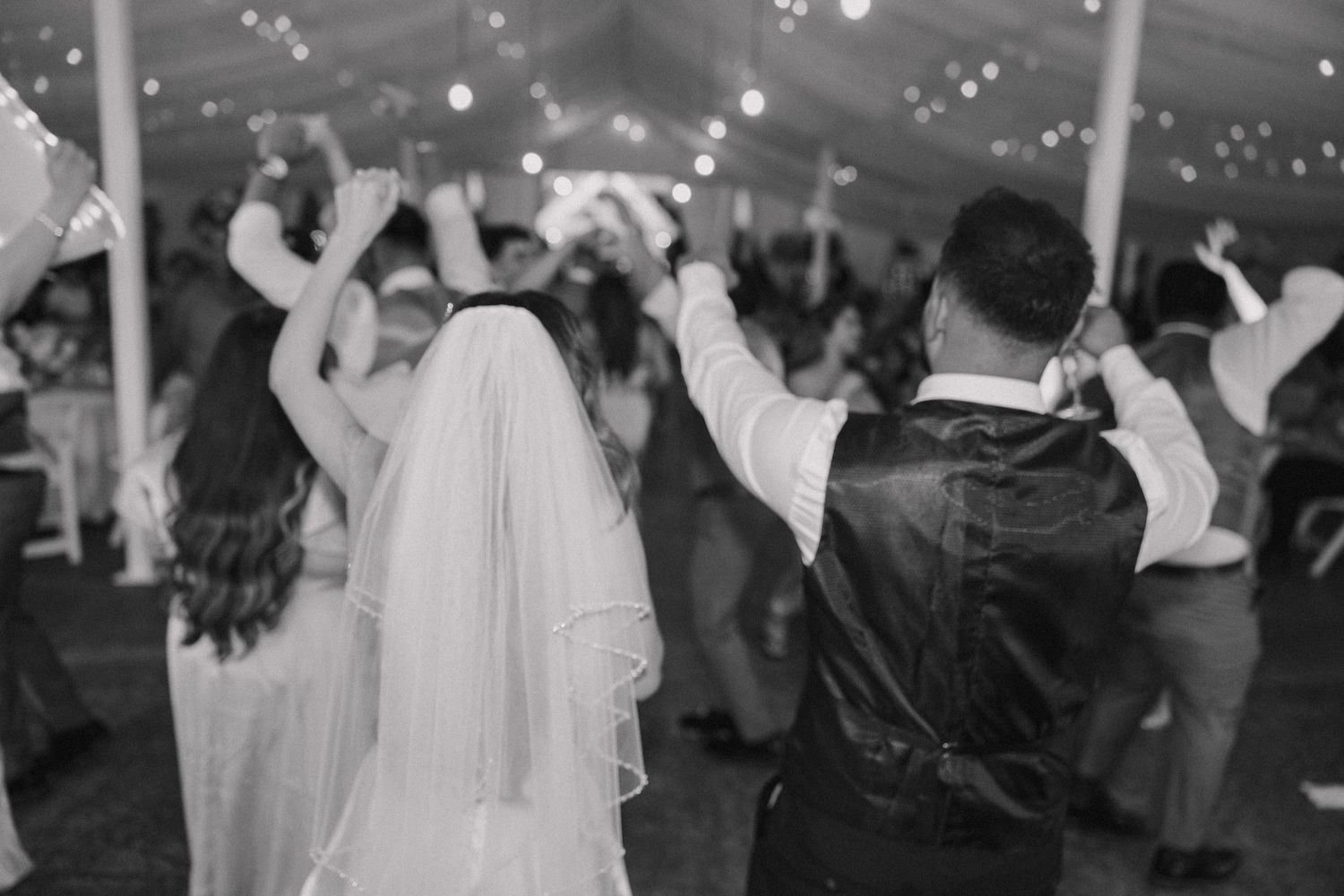 Black and white photo of bride and groom dancing with guests at elegant tented wedding reception.