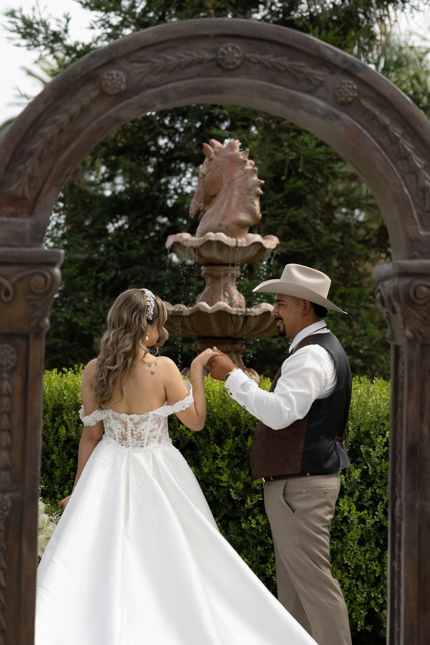 Bride and groom dancing under ornate arch with fountain, groom wearing cowboy hat and vest at outdoor wedding.