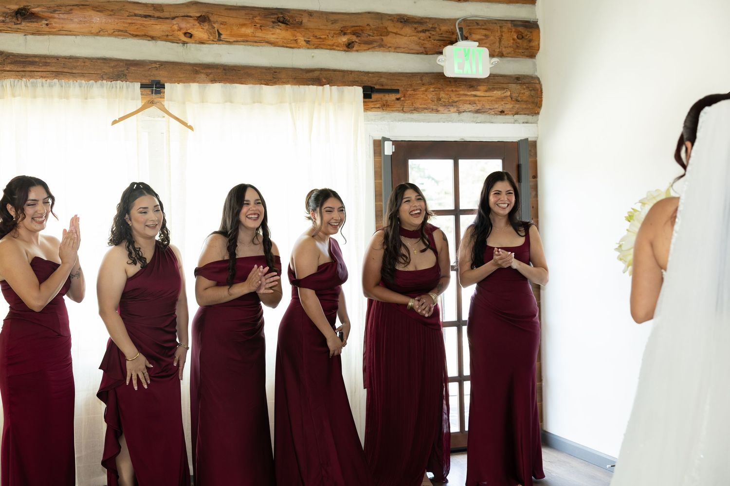 Bridesmaids in burgundy gowns react with joy as bride reveals her wedding dress in a rustic venue.