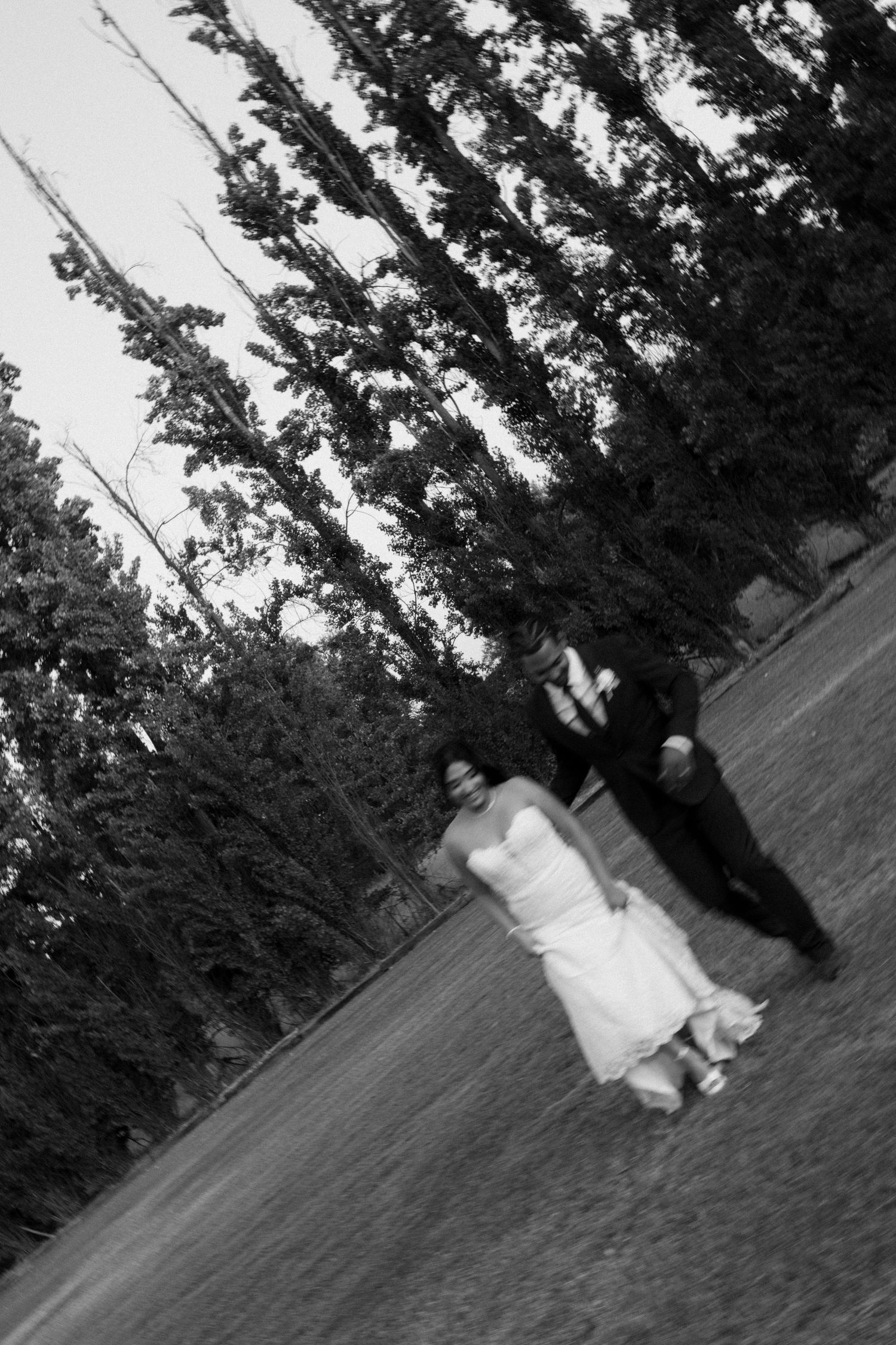 Black and white photo of bride and groom walking hand in hand across lawn near trees.