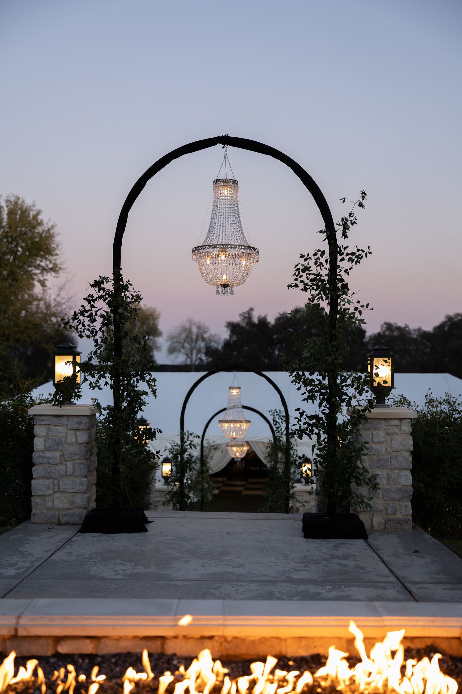 Elegant outdoor wedding arch with crystal chandelier and fire feature at dusk.