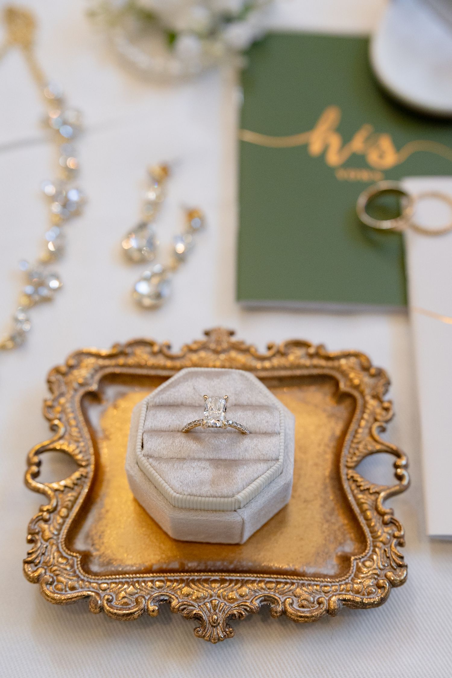 Diamond engagement ring in velvet box on ornate gold tray with crystal earrings and wedding bands nearby.