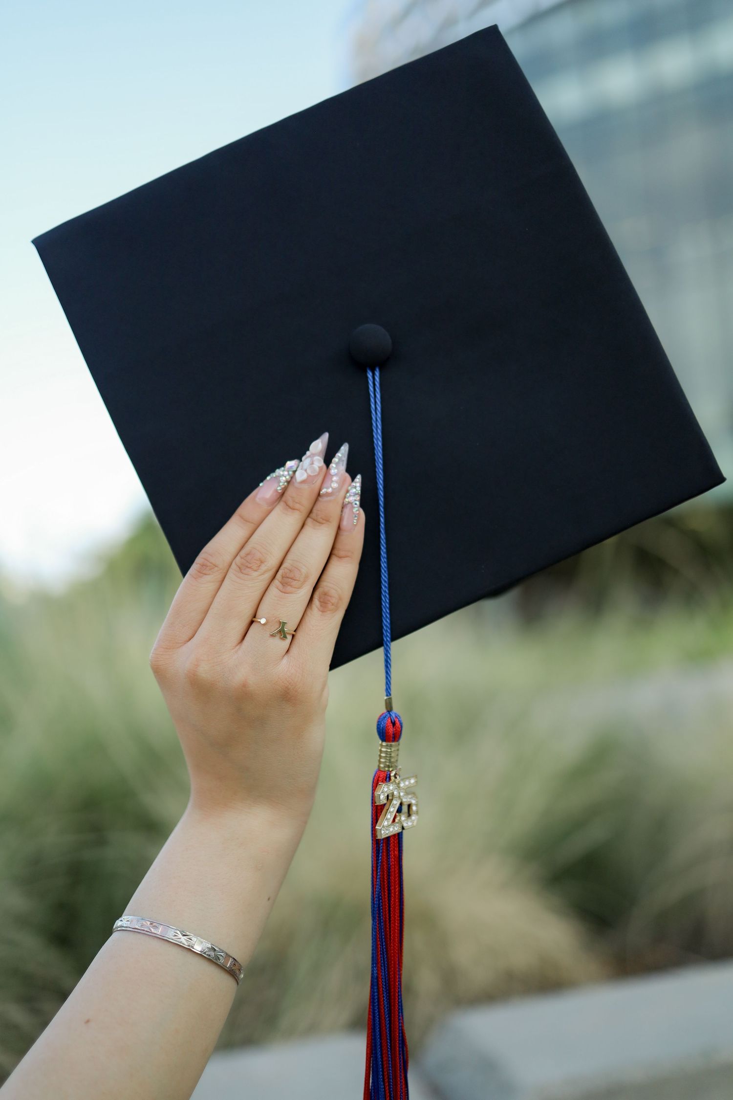 A hand wearing a ring holds a graduation cap with red and blue tassel against a blurred outdoor background.