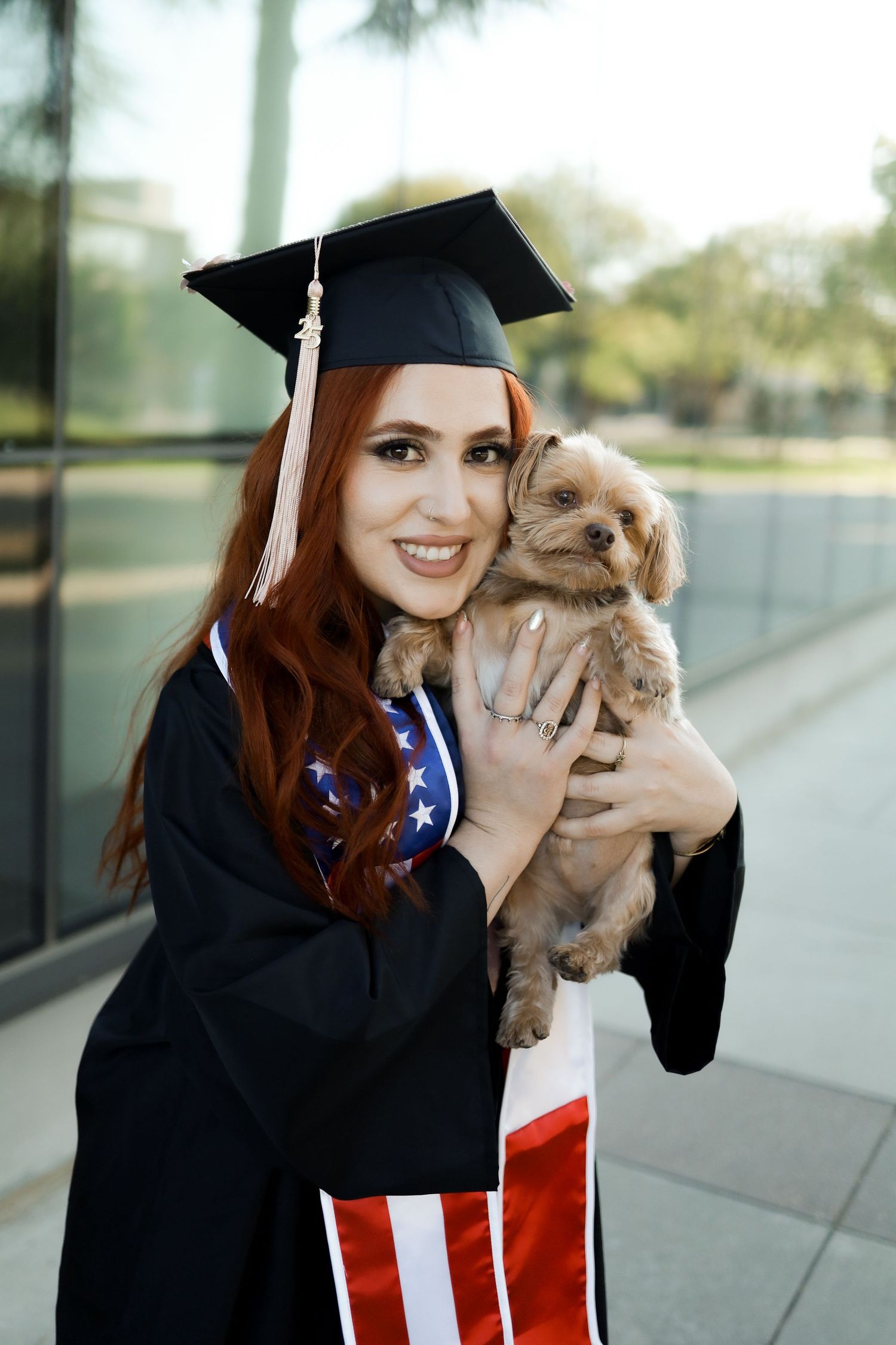 Graduate in black cap and gown holds small tan puppy while posing for graduation photos outdoors.