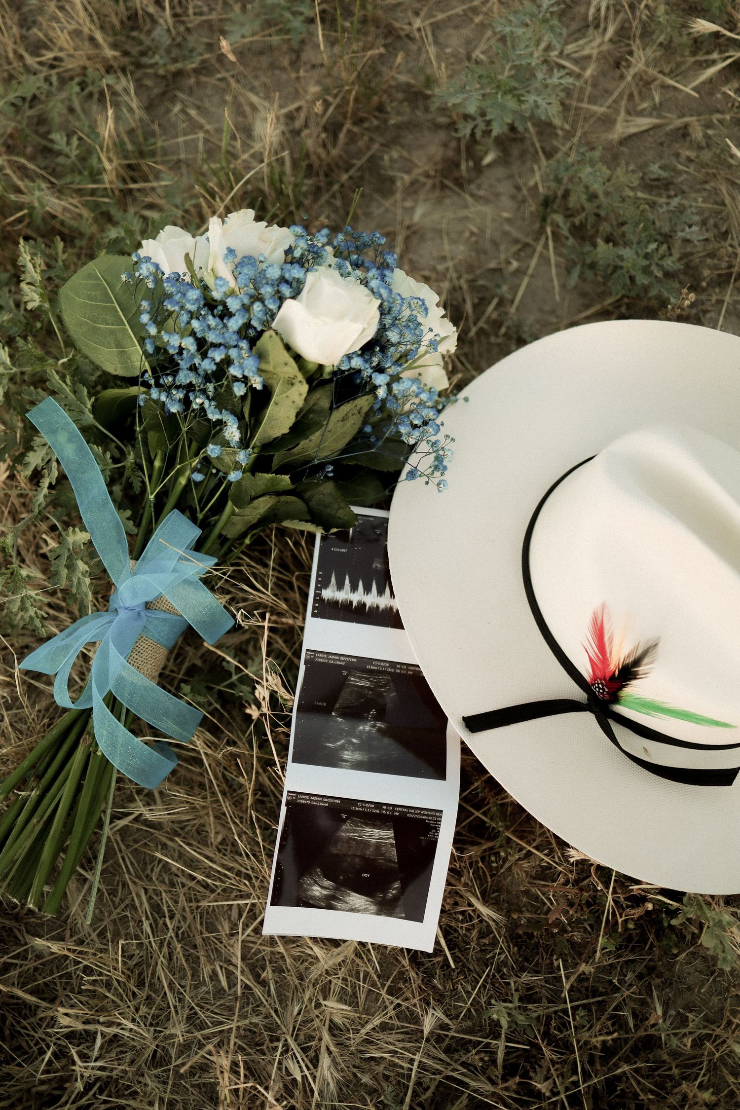 A white sun hat adorned with blue and white flowers and a black ribbon rests on grass with a Polaroid photo nearby.
