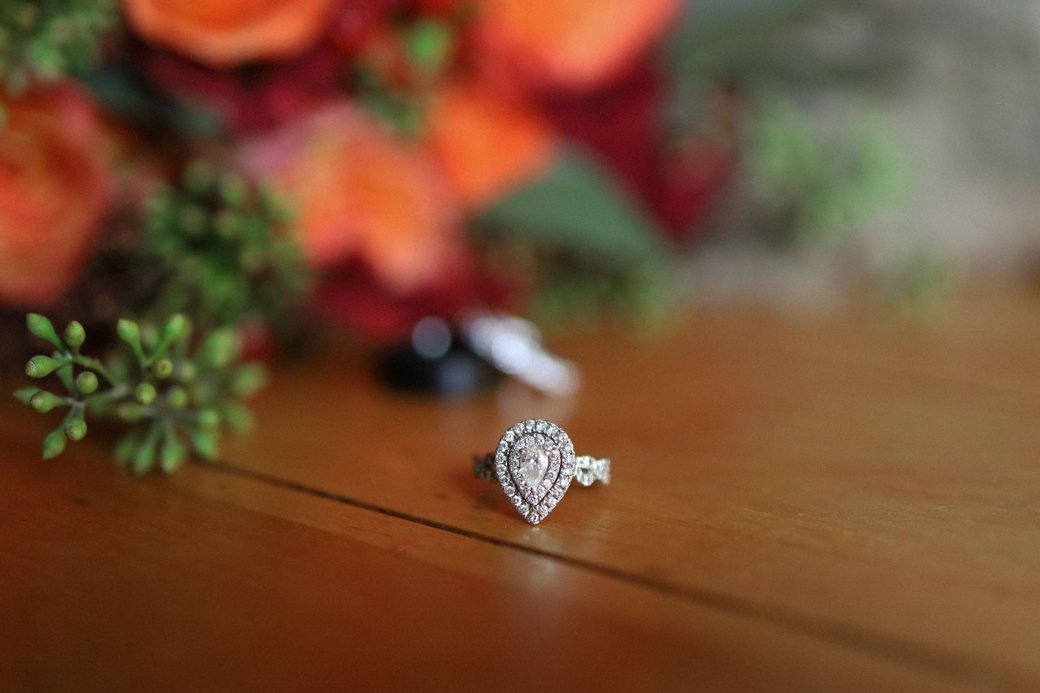 A sparkling pear-shaped diamond engagement ring on a wooden surface with orange flowers in background.