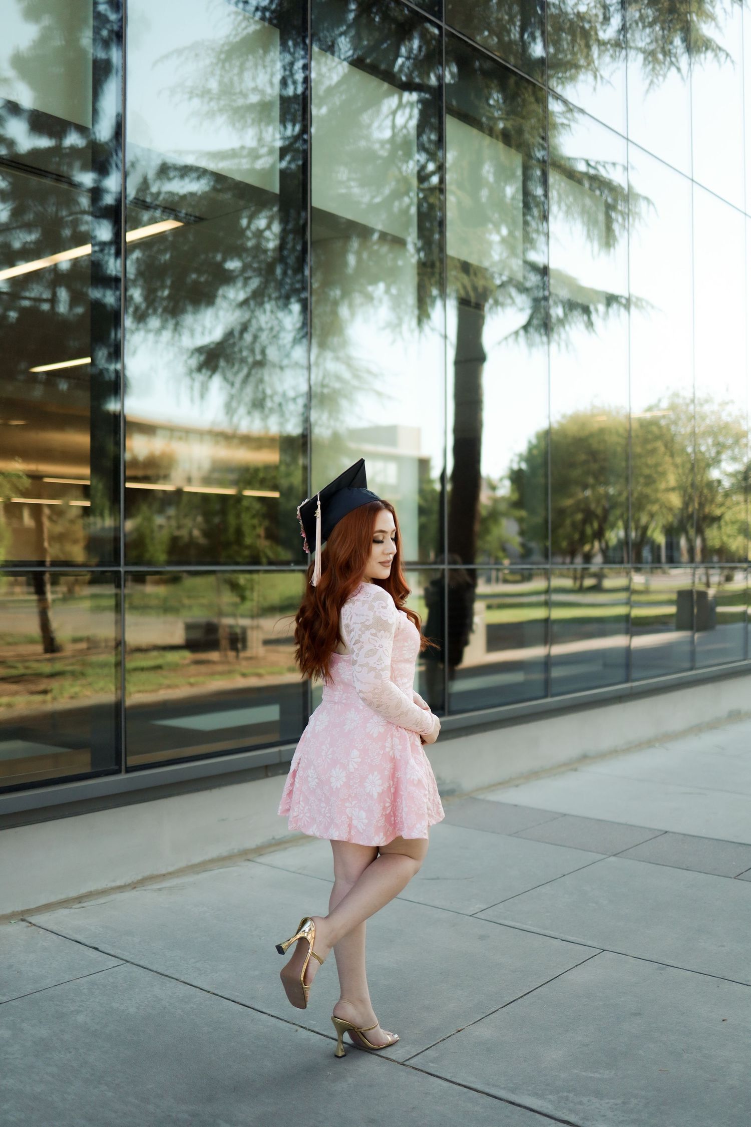 Graduate in pink lace dress and cap poses playfully outside modern glass building on sunny day.