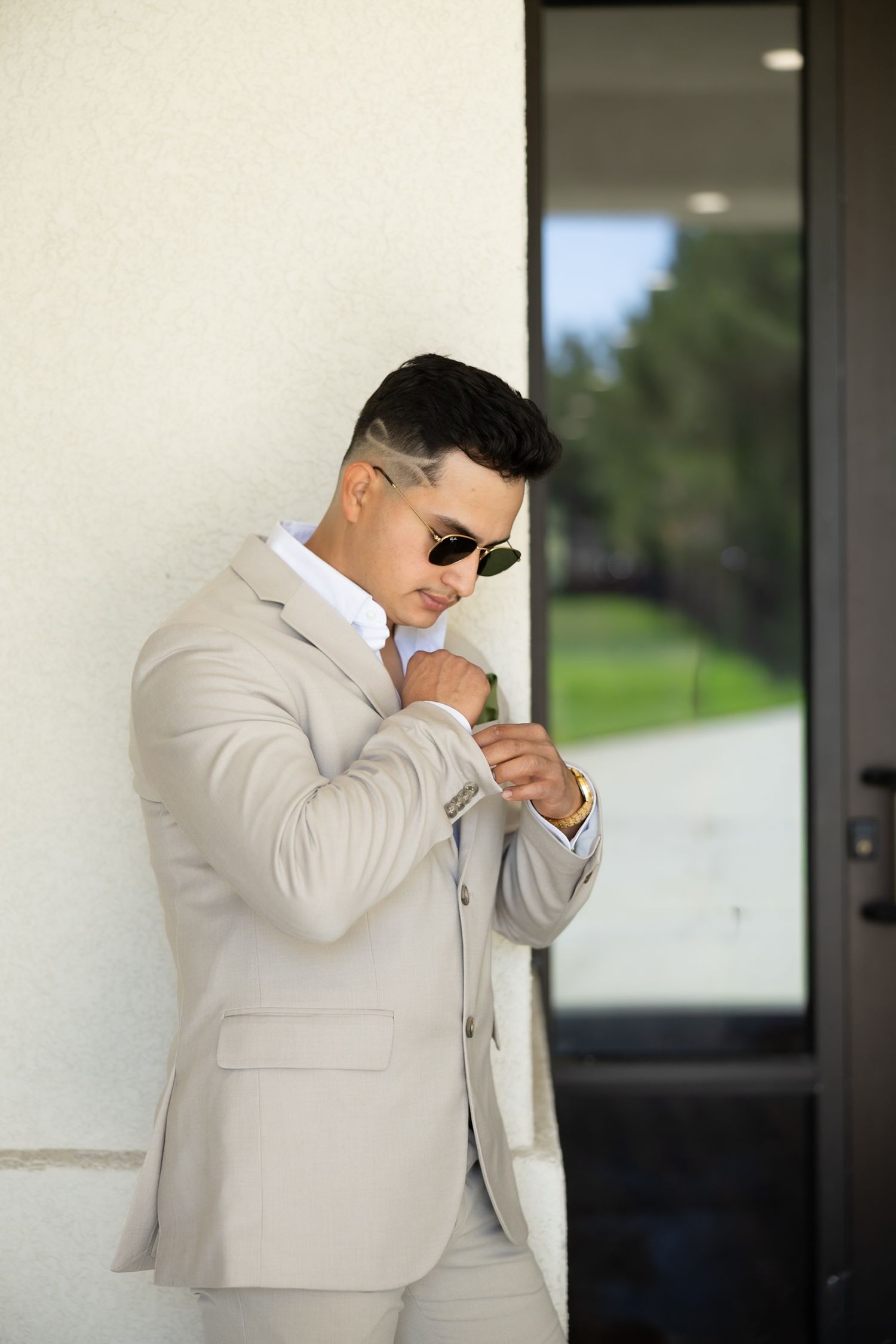 A stylish man in a beige suit and sunglasses adjusts his cufflinks while leaning against a white wall outdoors.
