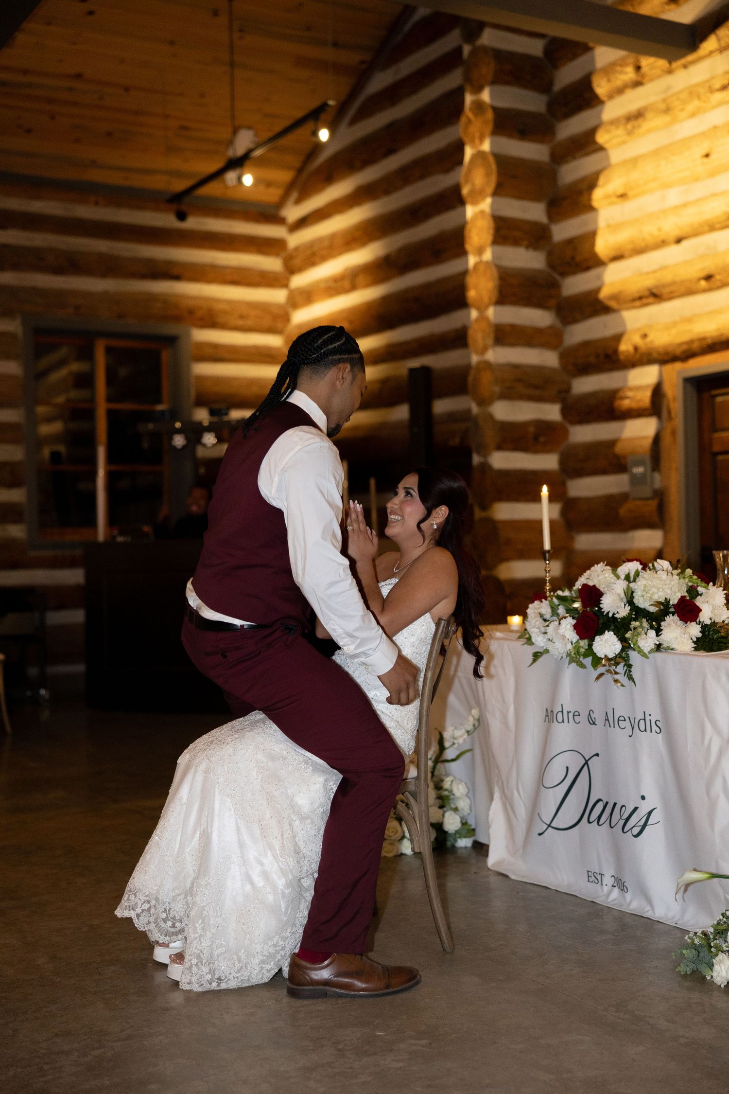 Bride and groom share playful intimate moment at sweetheart table inside rustic log cabin venue.