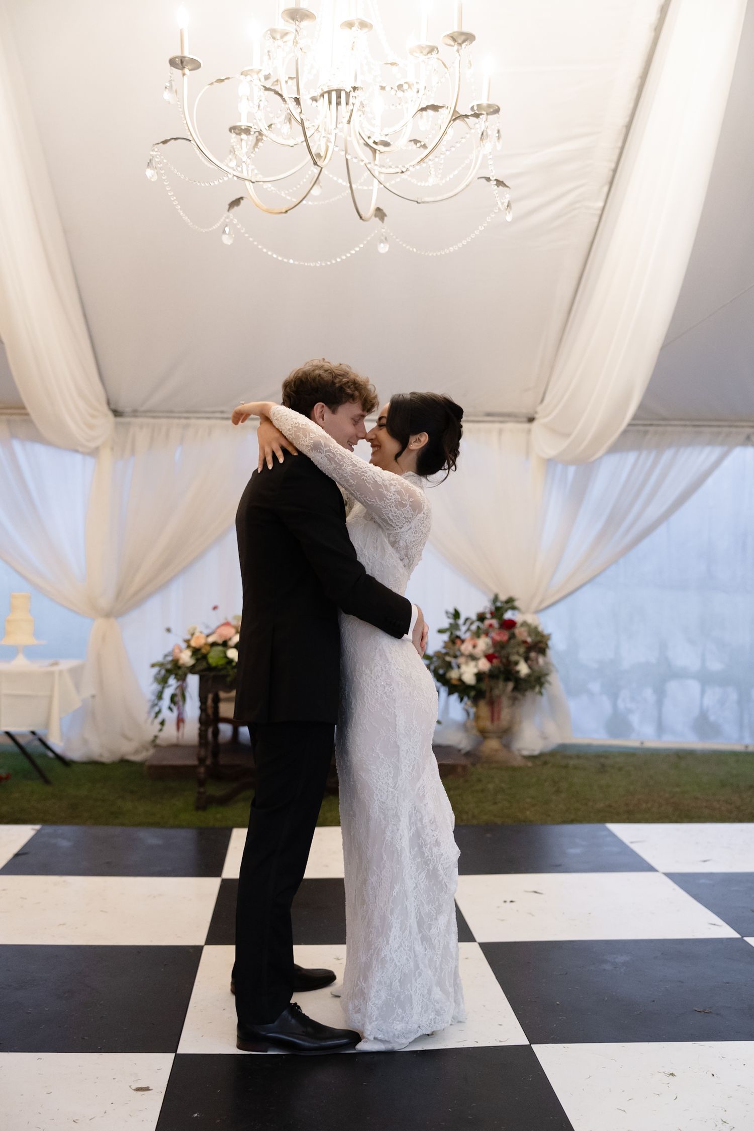 Bride and groom share first dance on checkered floor under chandelier in elegant white draped wedding tent.