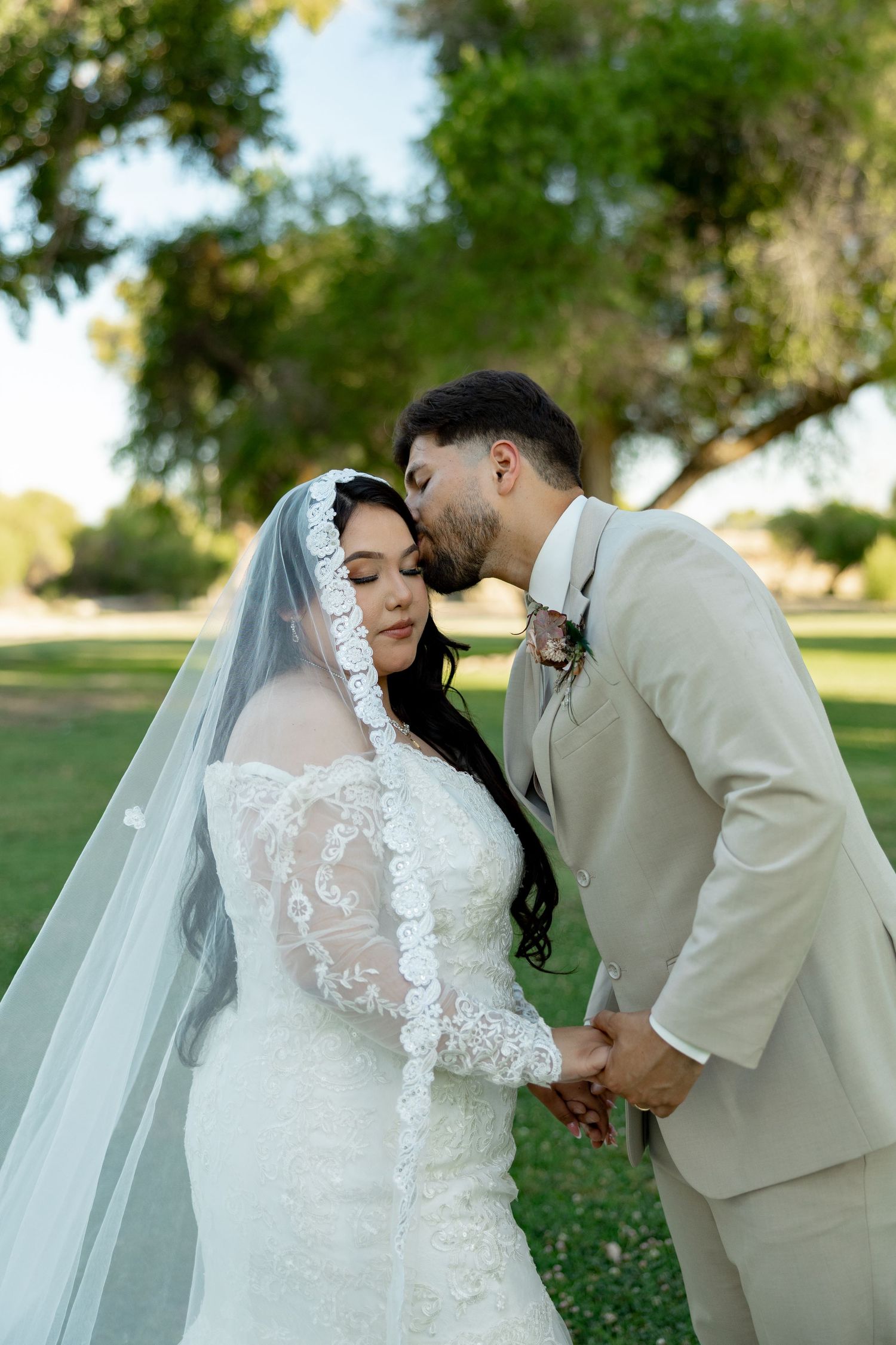 Groom in beige suit kisses bride wearing lace wedding dress and veil in a lush green park setting.