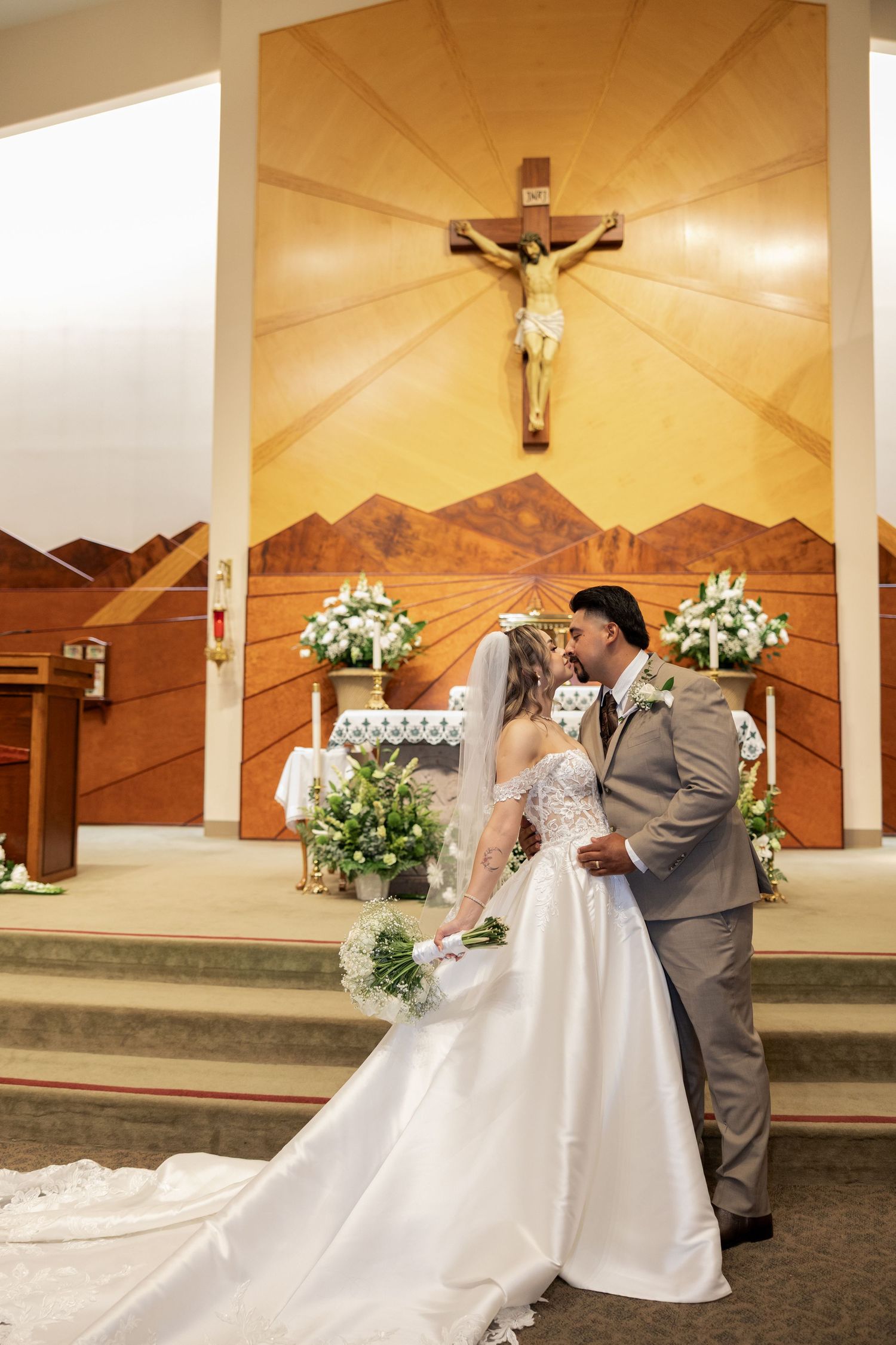 Bride and groom share first kiss at church altar with crucifix backdrop and white floral arrangements.