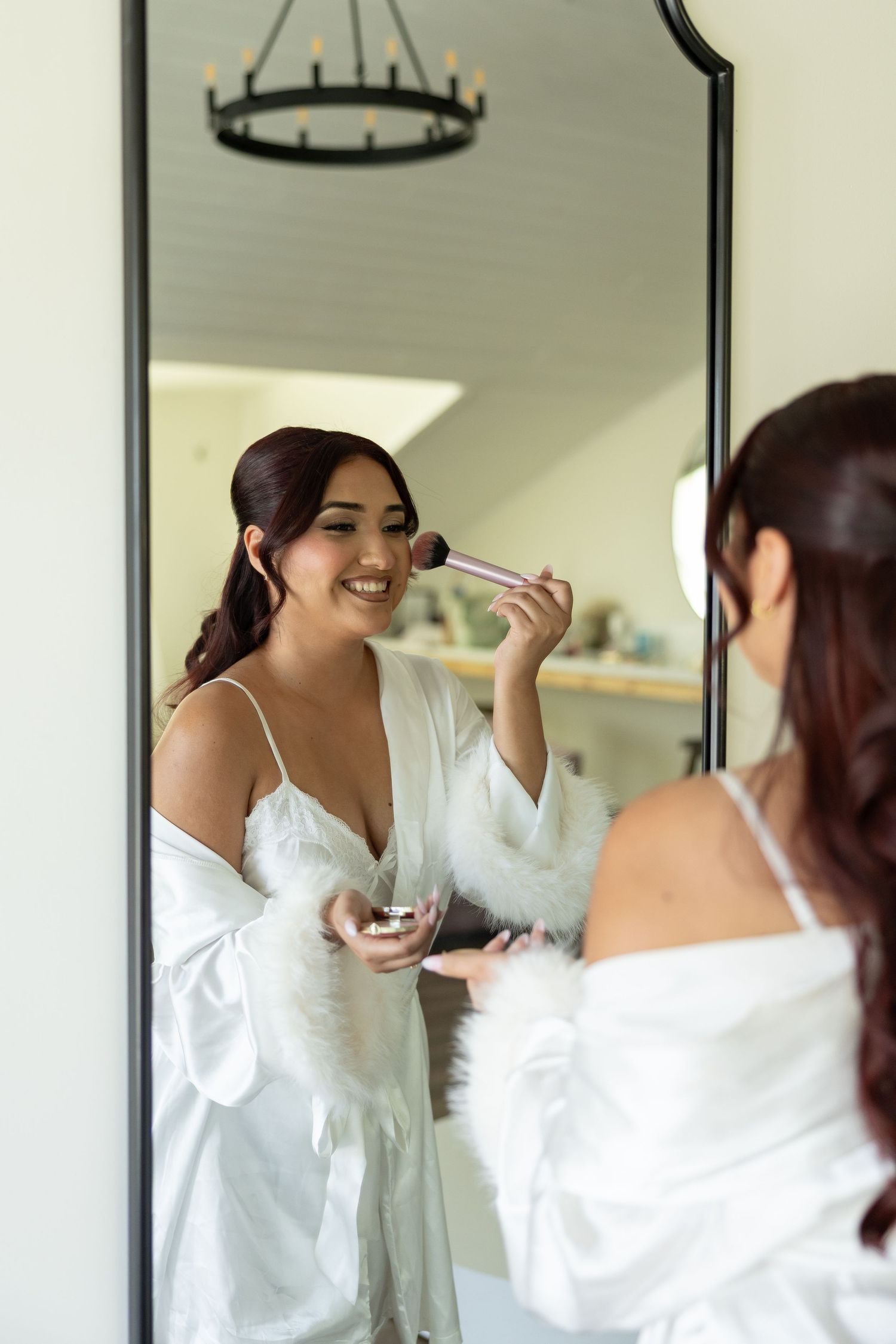 Smiling bride in white robe applies blush with makeup brush while getting ready in front of mirror.