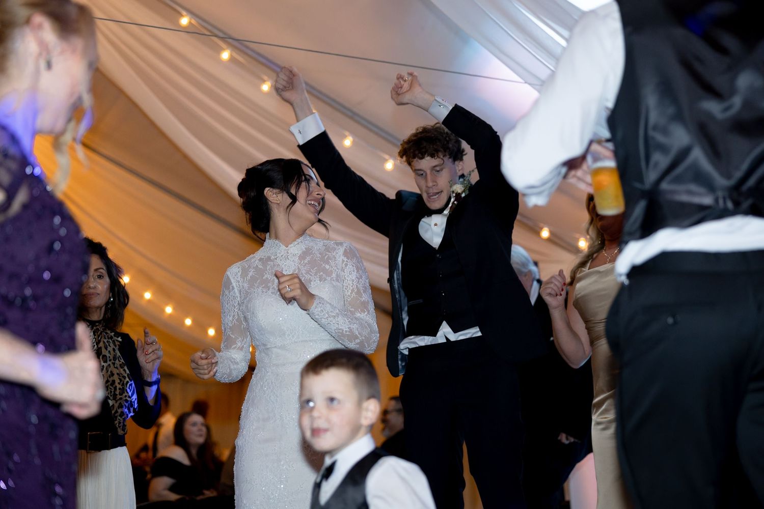 Bride and groom dancing joyfully with guests at a festive indoor wedding reception under string lights.