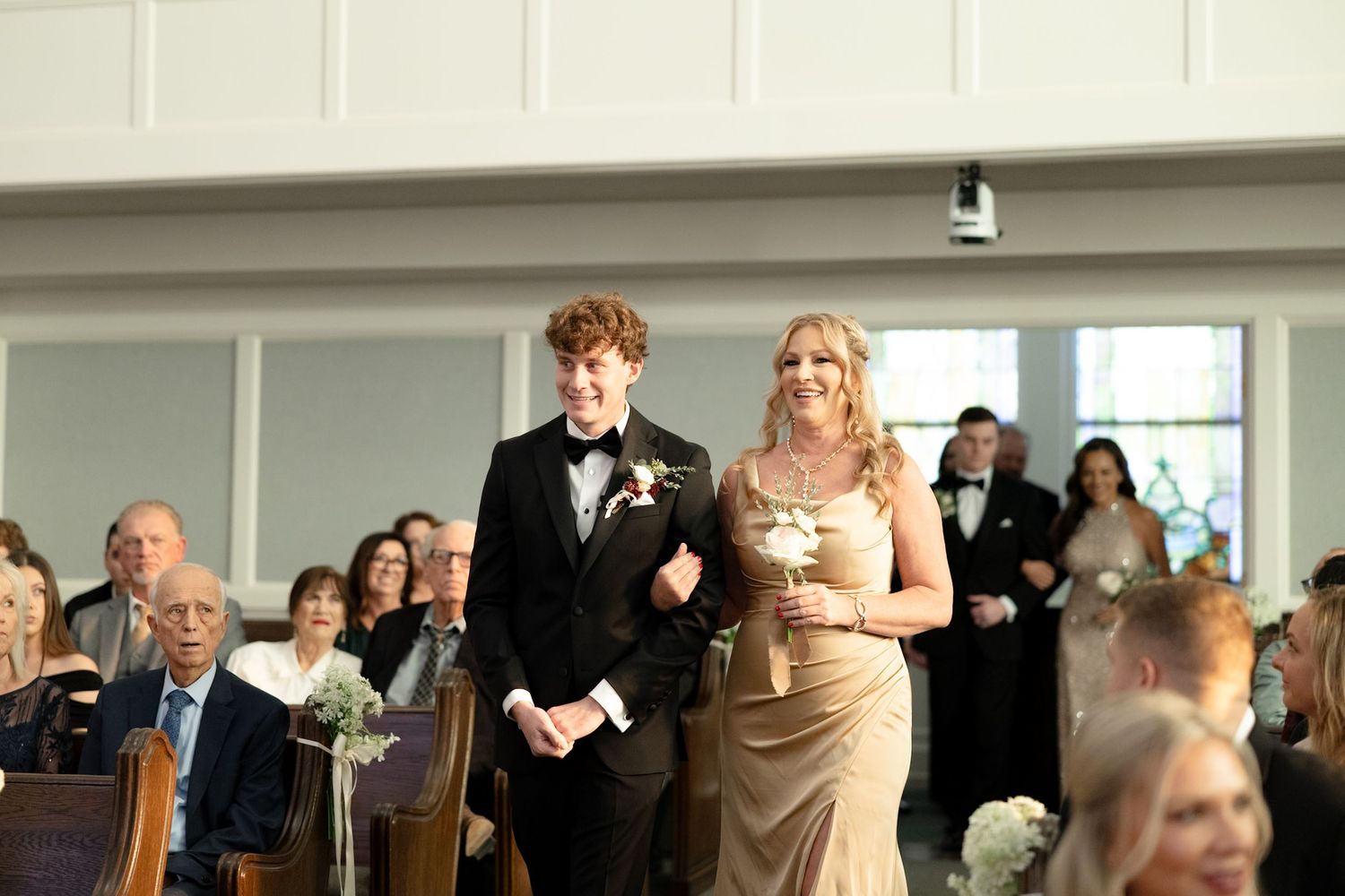 A groomsman in a black tuxedo escorts a bridesmaid in a gold dress down the church aisle at a wedding.